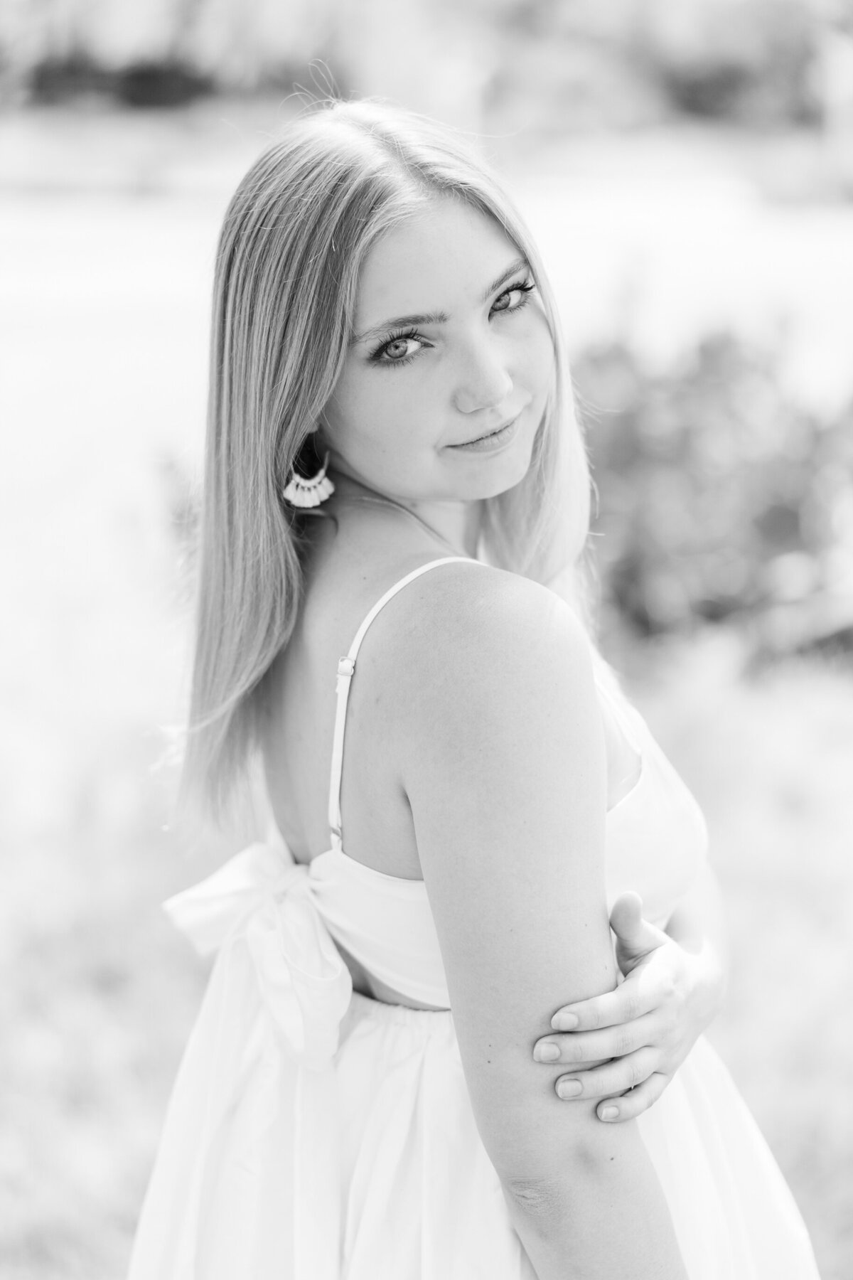 Black and white senior portrait of a young woman in a white dress with a bow detail, softly smiling over her shoulder. Photographed outdoors in natural light in downtown Fort Worth, Texas, with a classic and elegant feel.