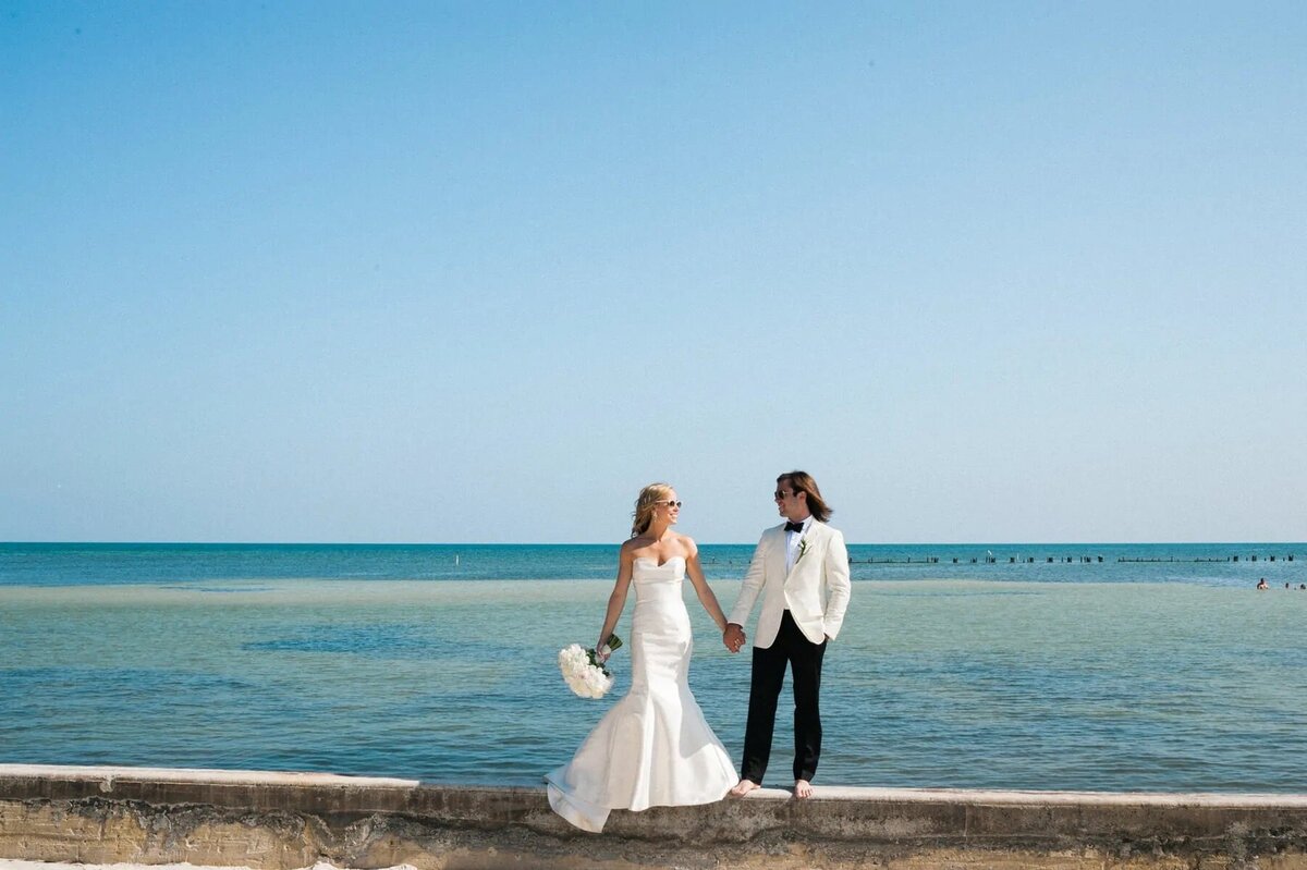 A couple stands hand in hand on a sea wall against the backdrop of a calm blue sea, under a clear sky