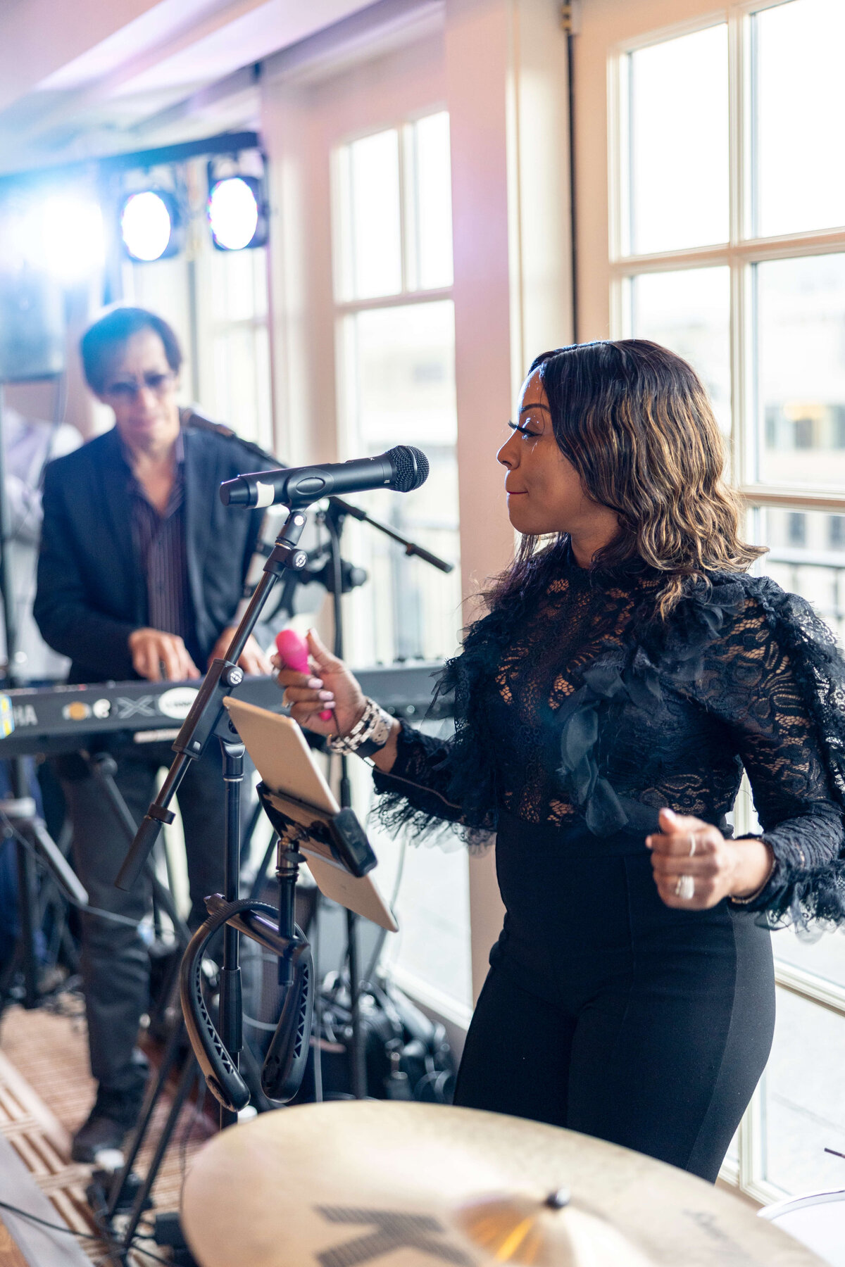 A woman sings into a microphone, wearing a black lace top and pants. A keyboard player is in the background with stage lights shining. Theyre performing in a well-lit indoor area with large windows.