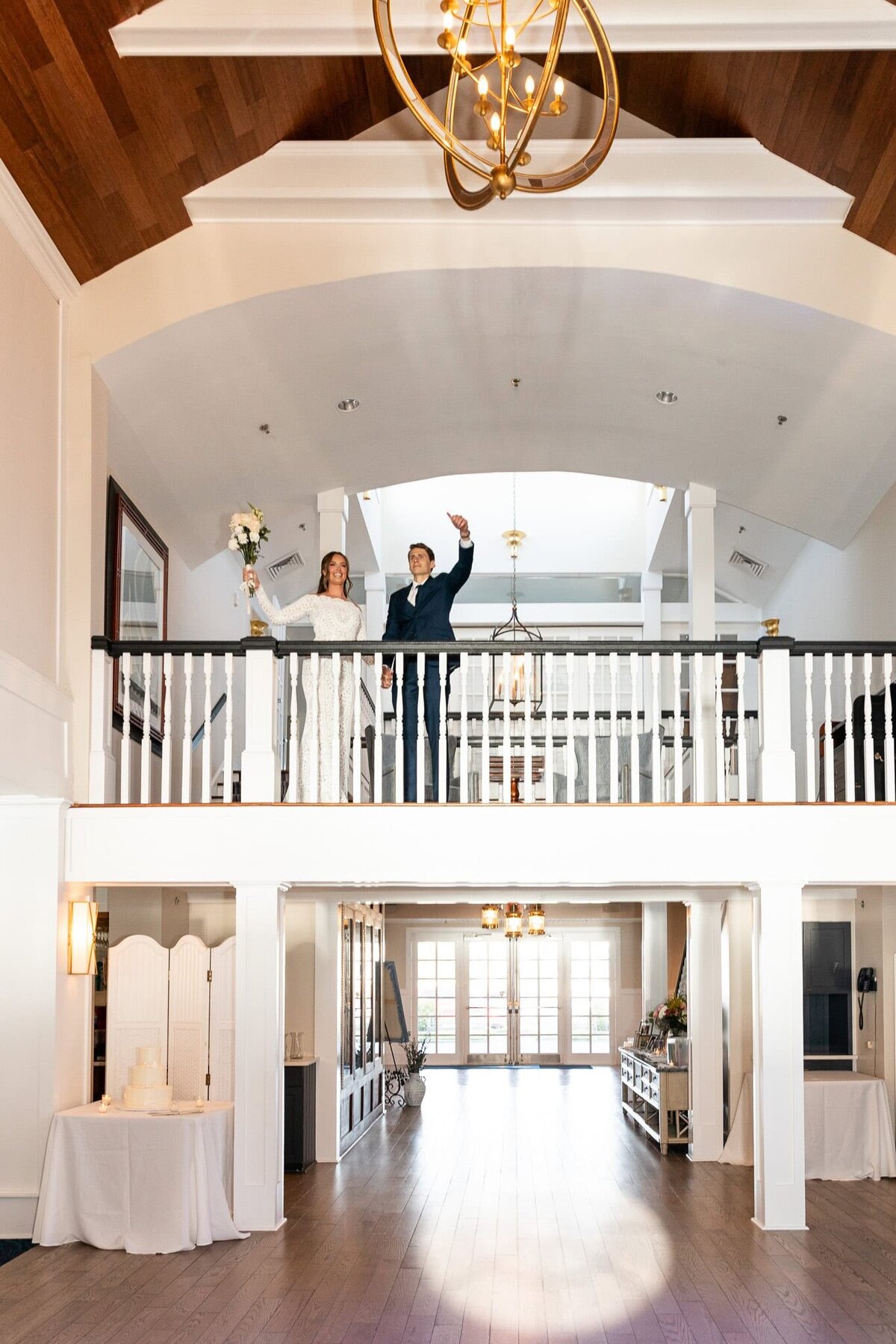 The newlyweds stand on a white-railed balcony, hands raised in celebration as they look down at their wedding guests below. The bride’s long-sleeved gown and the groom’s navy suit create a striking contrast against the airy, light-filled venue, with a golden chandelier casting a warm glow over the moment