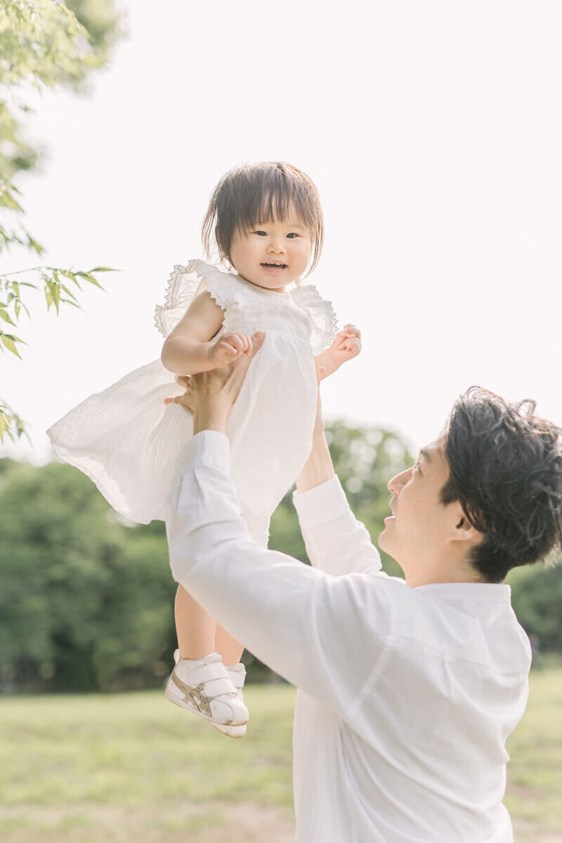 Father holding his daughter up in a park located in Shibuya, Japan. The girl 's white dress is moving in the wind.