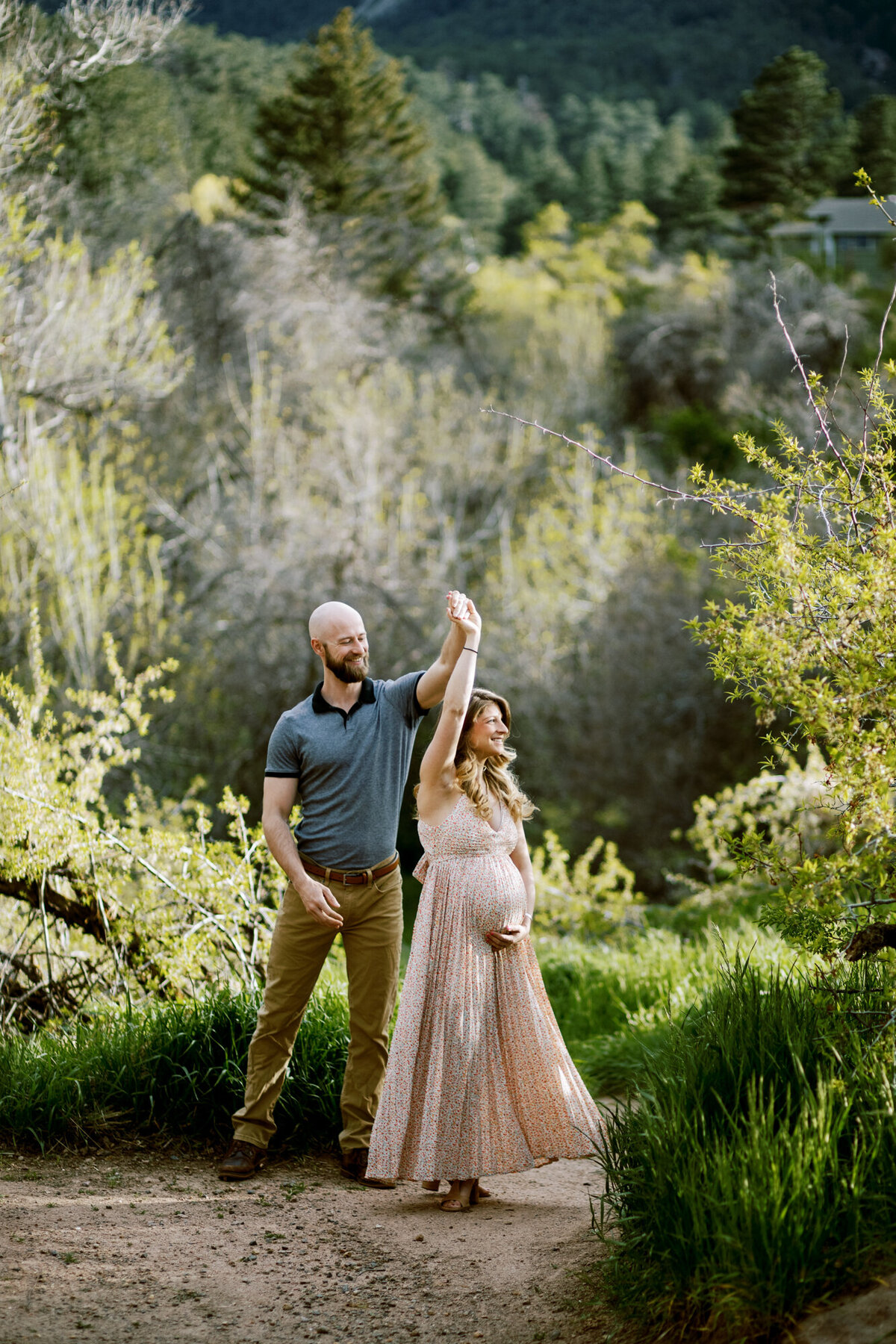 a man spins his pregnant wife around while she clutches her baby bump and wears a pink sun dress at a park in boulder, colorado