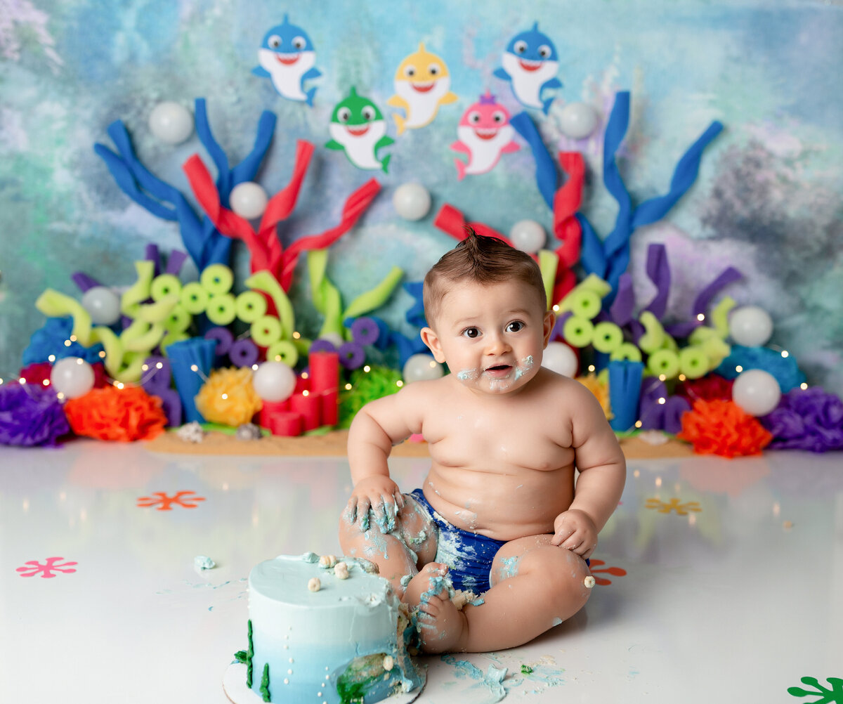 Baby Shark themed cake smash at West Palm Beach and Lake Worth, FL cake smash photographer. Baby is sitting behind underwater-themed cake with his legs covered in icing. In the background, there is coral and the baby shark family.