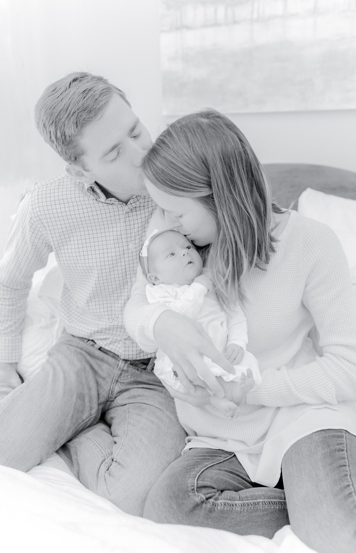 Black and white newborn family photography, parents cuddling baby, Birmingham newborn photographer – A heartfelt moment as a mother kisses her newborn while the father lovingly kisses her forehead, capturing the warmth and tenderness of early parenthood.