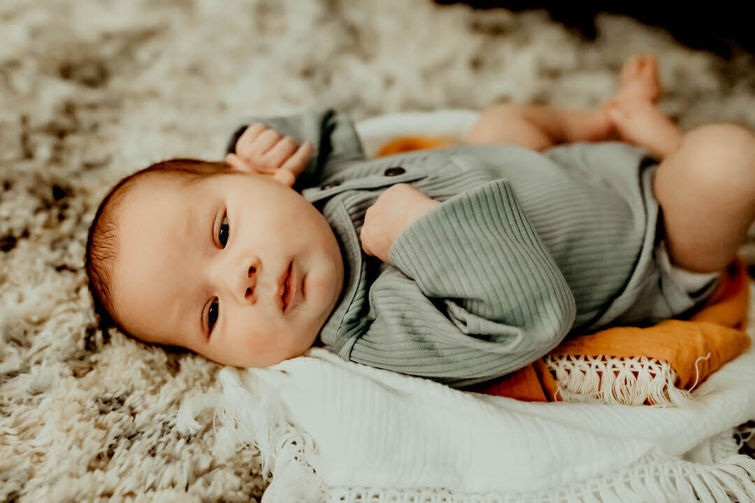 A baby in a gray outfit lies on a soft blanket on a textured rug, looking towards the camera.