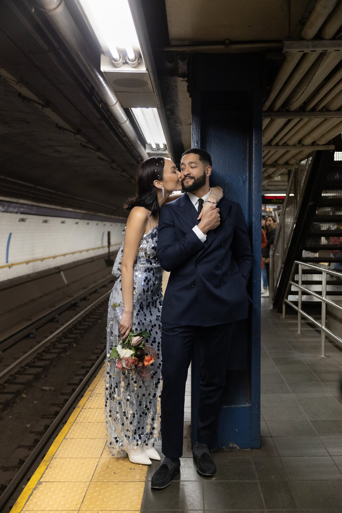 The couple shares a playful laugh on the subway platform, surrounded by the urban energy of New York City.