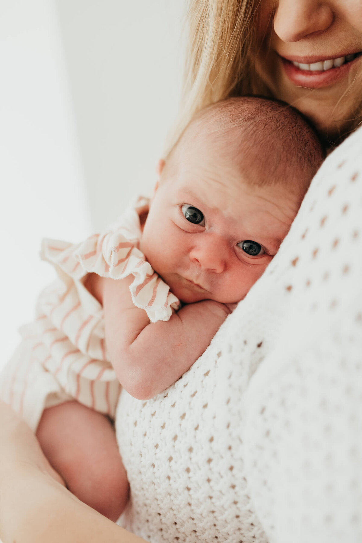 A newborn baby girl looks directly at the camera while wearing a cute white and pink striped romper, her mother is holding her and smiling