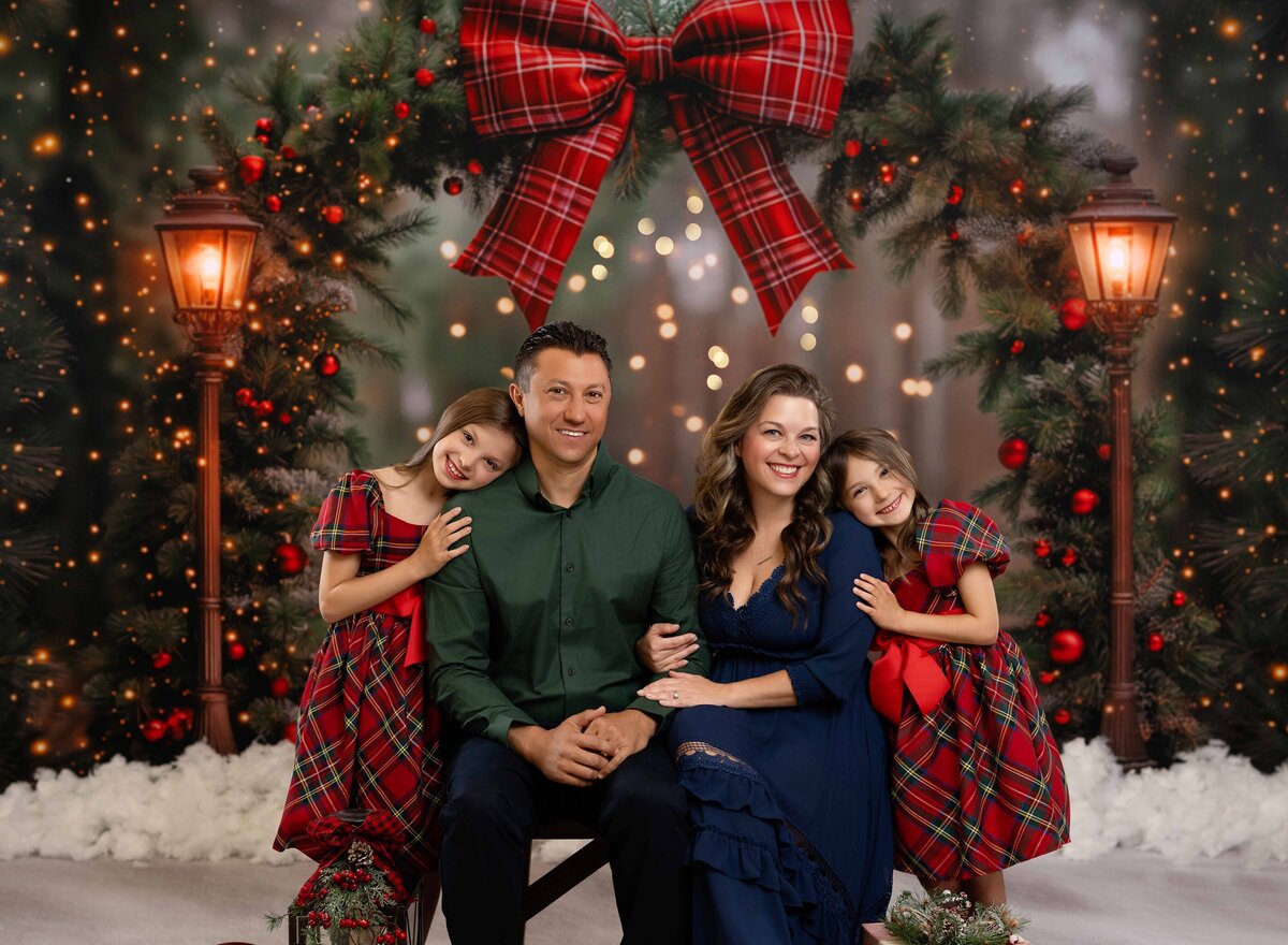 Family of 4 sitting on a bench in front of a Christmas Arch and smiling at the camera.