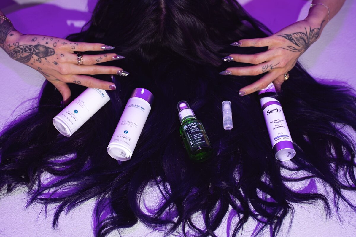 Woman with long dark hair and tattoos has several products displayed in a flat lay across her hair on a white surface.