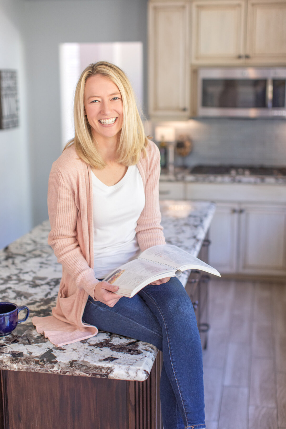 A cheerful woman in a soft pink sweater smiles while seated on a kitchen counter, holding a cookbook—captured to reflect lifestyle branding for wellness coaches and content creators.