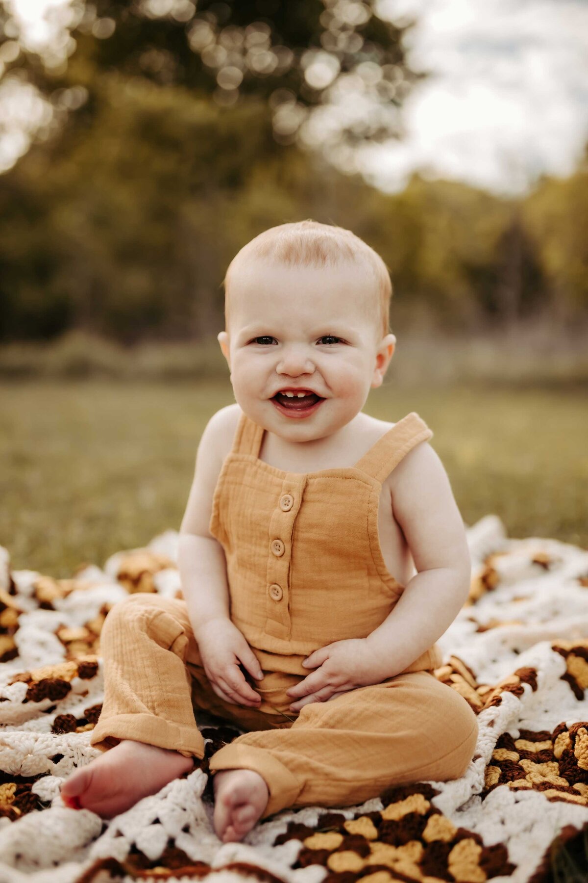 young boy sitting on a macrame blanket smiling. He is wearing yellow overalls