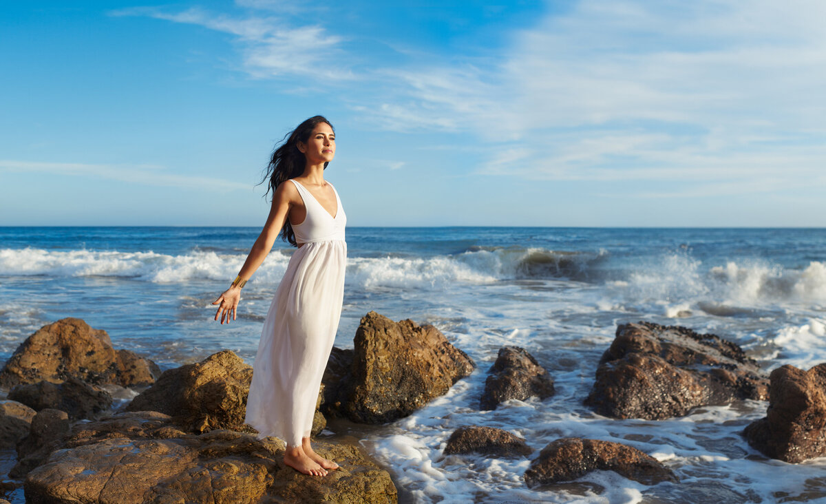 Elegant portrait of Kimberly Snyder standing by the ocean, dressed in a flowing white dress, captured by Ylva Erevall.