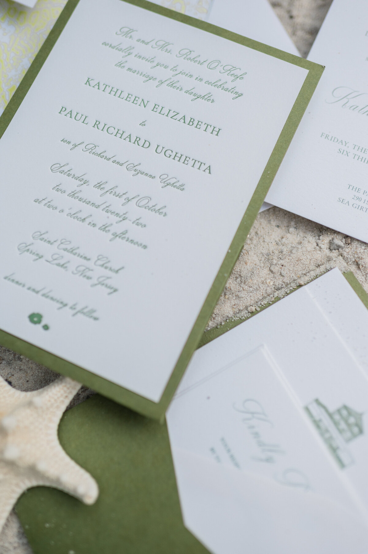A close-up of a wedding invitation set with green borders, placed on a sandy surface with a starfish beside it. The invitation announces the wedding of Kathleen Elizabeth and Paul Richard Ughetta at Saint Catharine Church in Spring Lake, New Jersey.