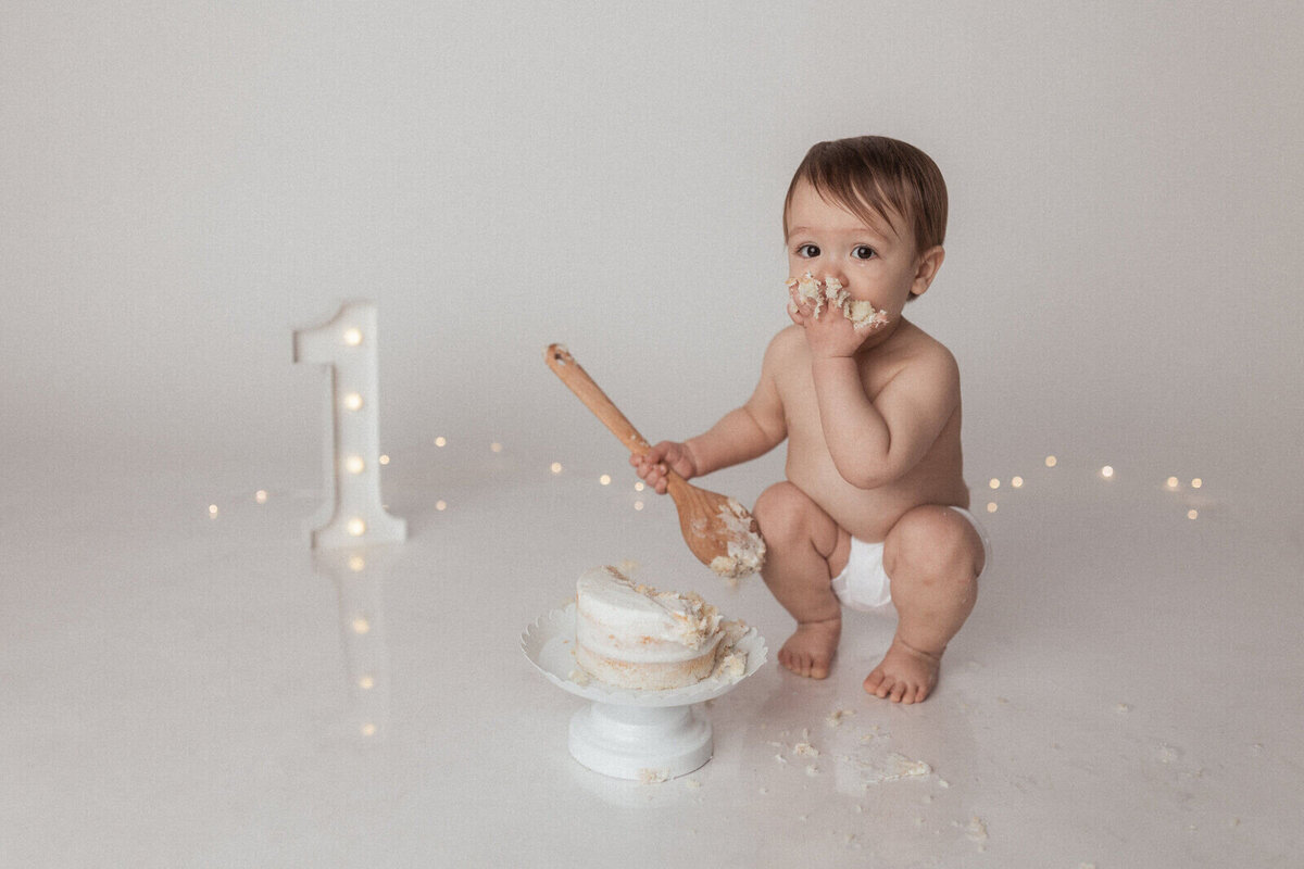 A baby boy wearing only a diaper squats next to a small white cake, eating cake with one hand while holding a wooden spoon in the other. A lit number one sign and fairy lights are in the background.