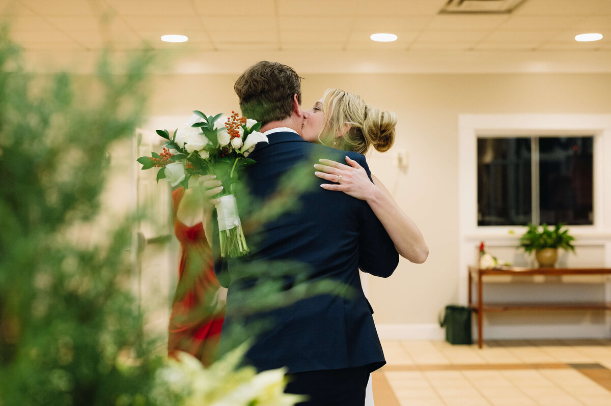 candid photo of bride kissing her grooms cheek as she holds his shoulder and with her other hand holds her wedding bouquet