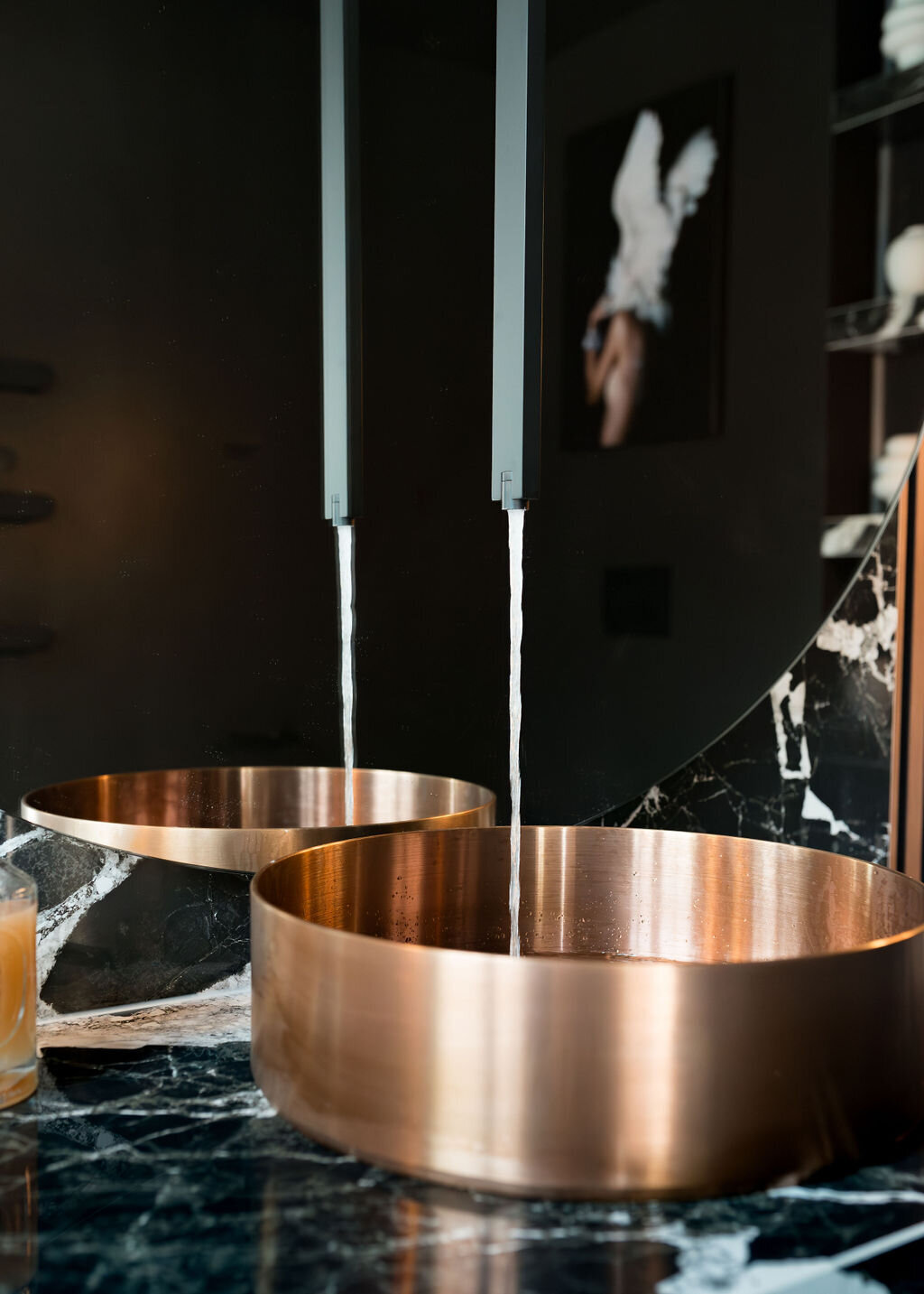 A modern bathroom sink features a round, metallic copper basin with water flowing from a stylish, minimalist matte black faucet. The countertop is made of black marble with white veining. Artwork hangs in the background, seen in the mirror.