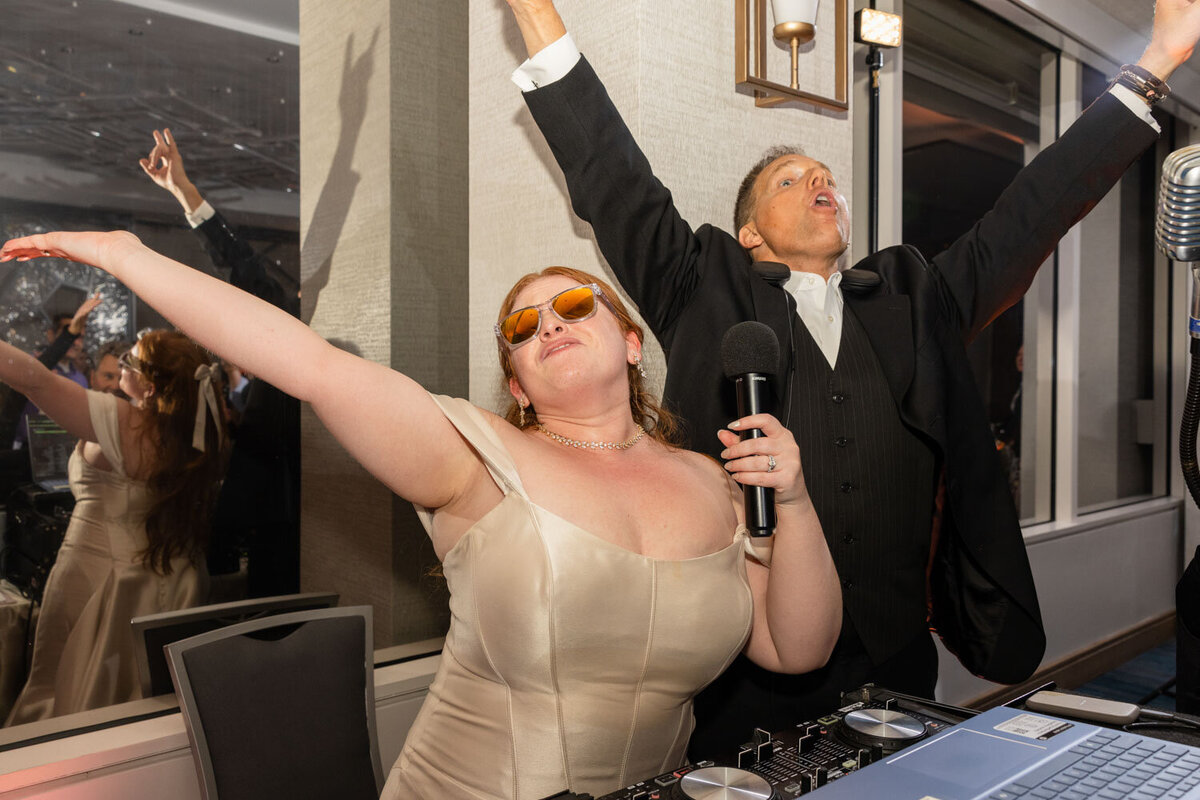 A man in a tuxedo holds a microphone while a woman in sunglasses holds her arms in the air