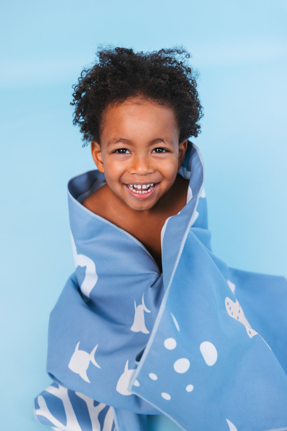 Smiling child wrapped in a blue Swim Essentials towel with playful sea animal prints, sitting on a matching blue background.