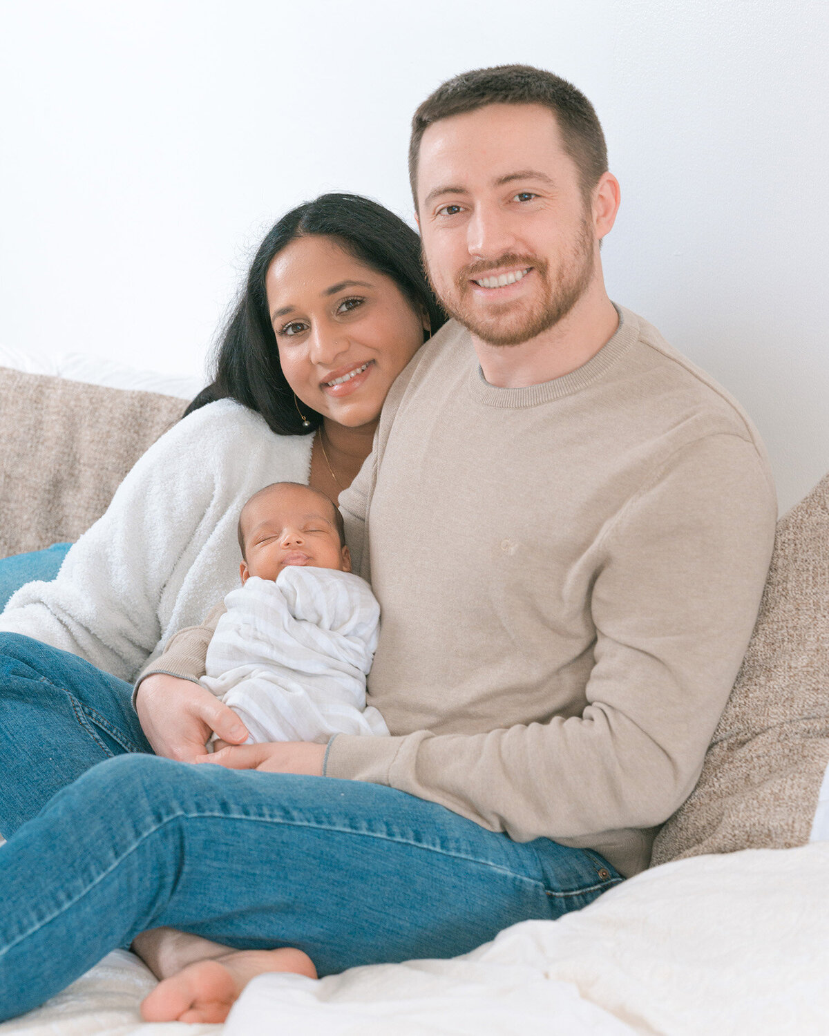 Indian mother and white father sitting on white bed holding newborn baby and smiling in natural light, captured by Cupertino newborn photographer during in-home session.