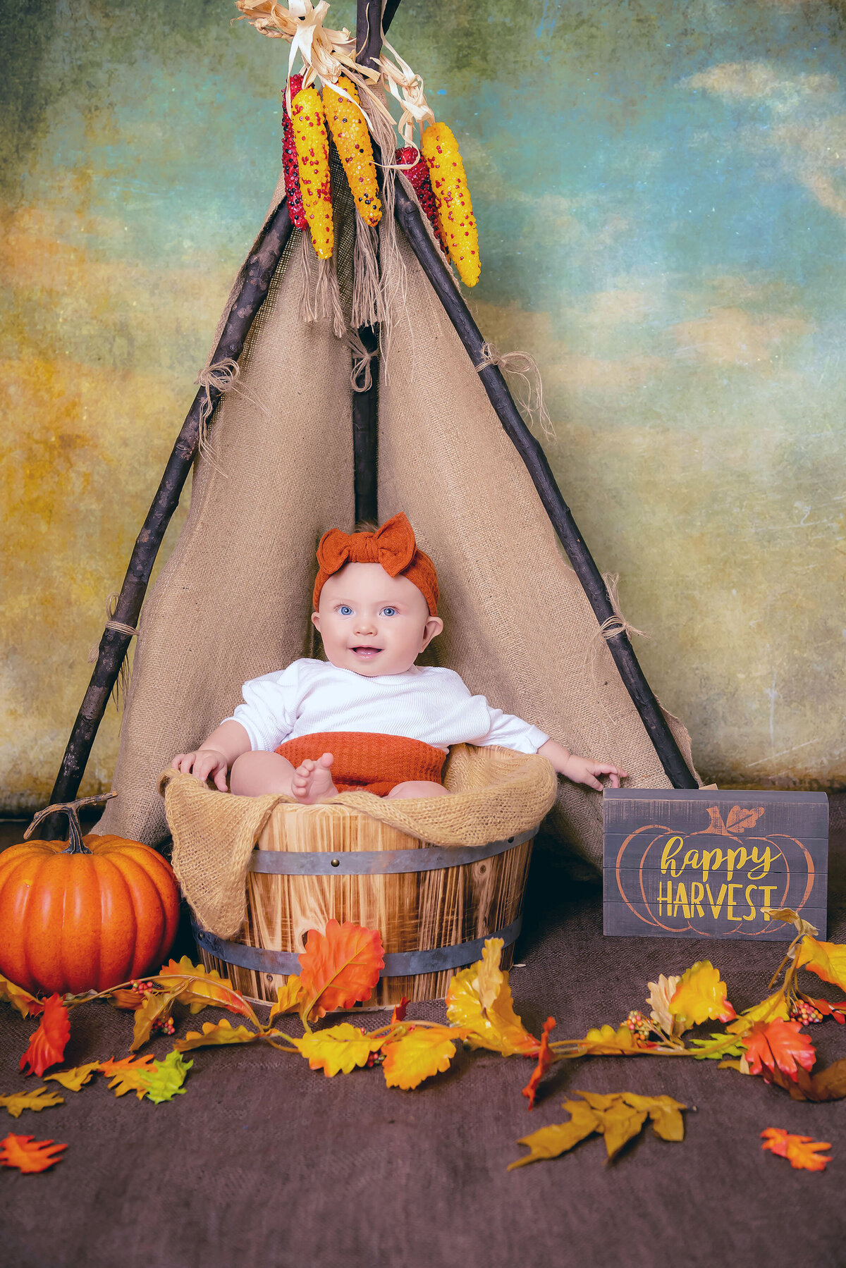 An Orange County milestone photography image of a baby sitting in a bucket in a fall themed studio smiling with an orange bow
