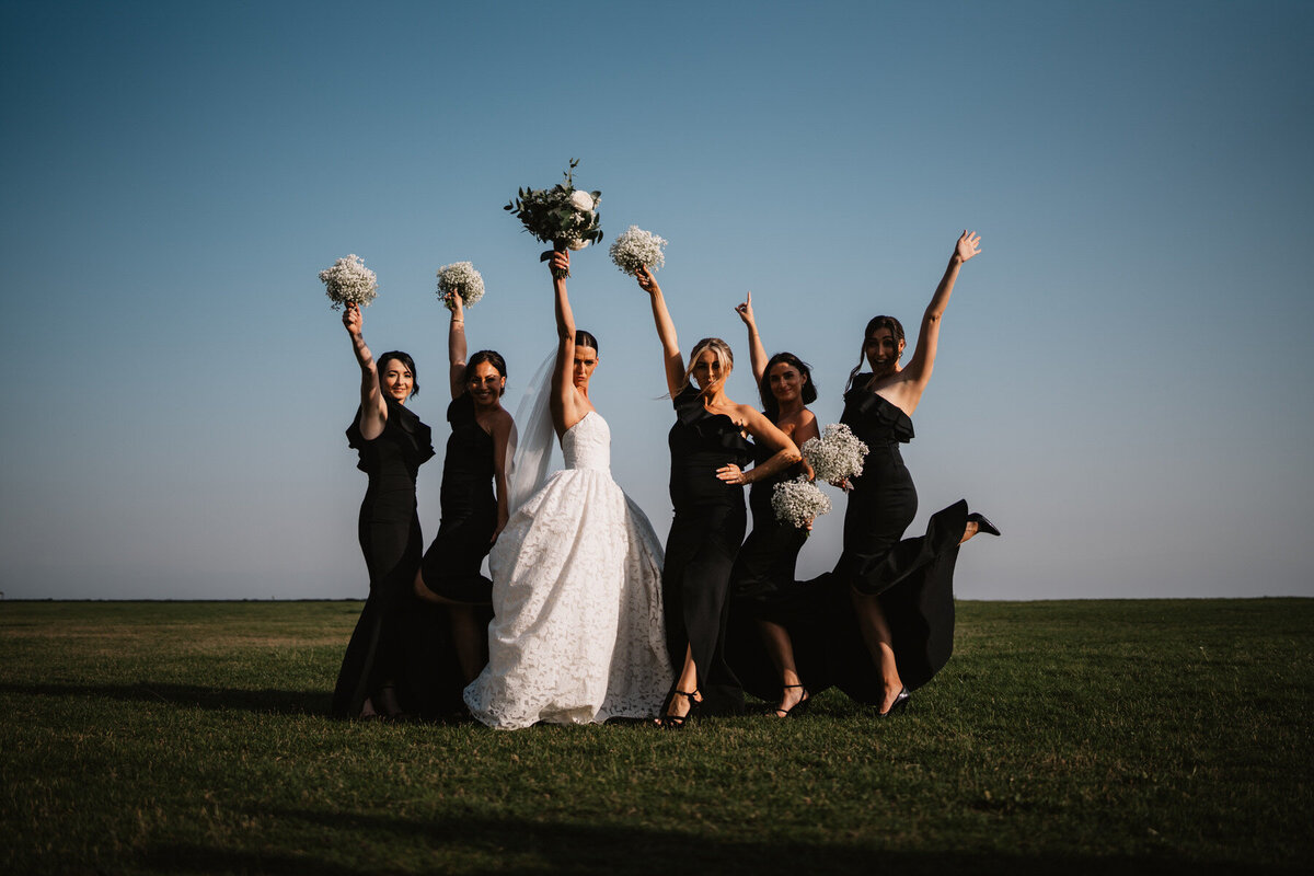 Outdoor wedding photo of bride with bridesmaids in black dresses holding bouquets in the air, Elegant wedding photography, Lytham wedding photography, Lancashire wedding photographer