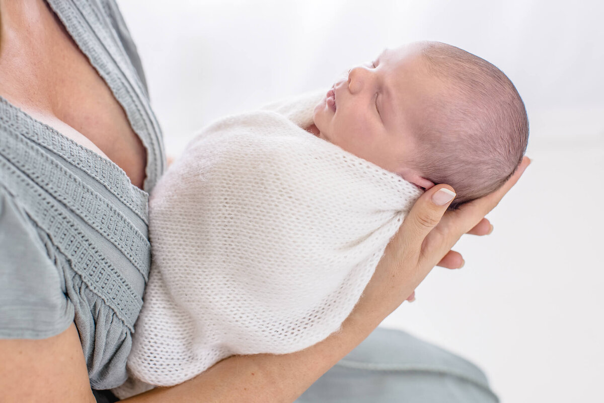 A newborn baby wrapped in a soft white blanket is cradled in the arms of an adult wearing a gray shirt. The scene is gentle and serene, with the baby peacefully sleeping. The background is softly blurred.