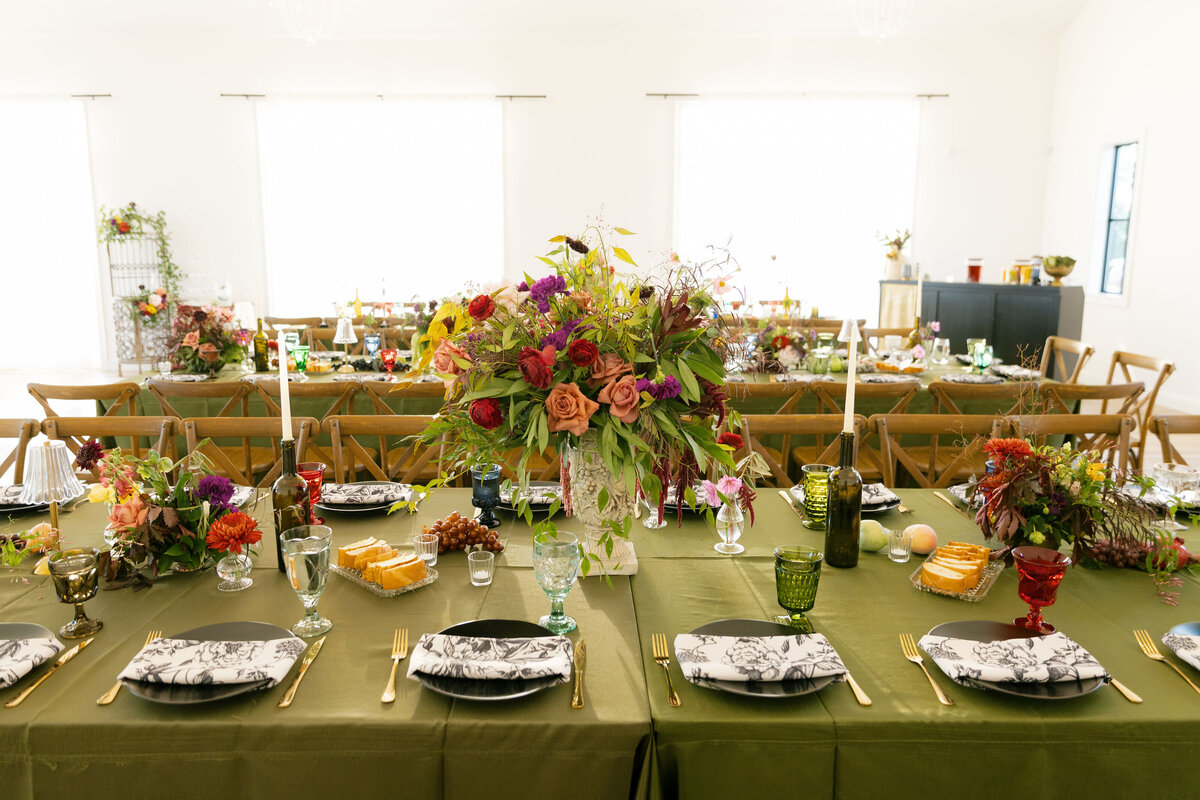 A long, greenery-covered wedding reception table styled with layered floral centerpieces featuring roses, garden blooms, vibrant colors, textured fruits, candles, and vintage-inspired glassware for an elevated wedding tablescape design by a floral stylist.