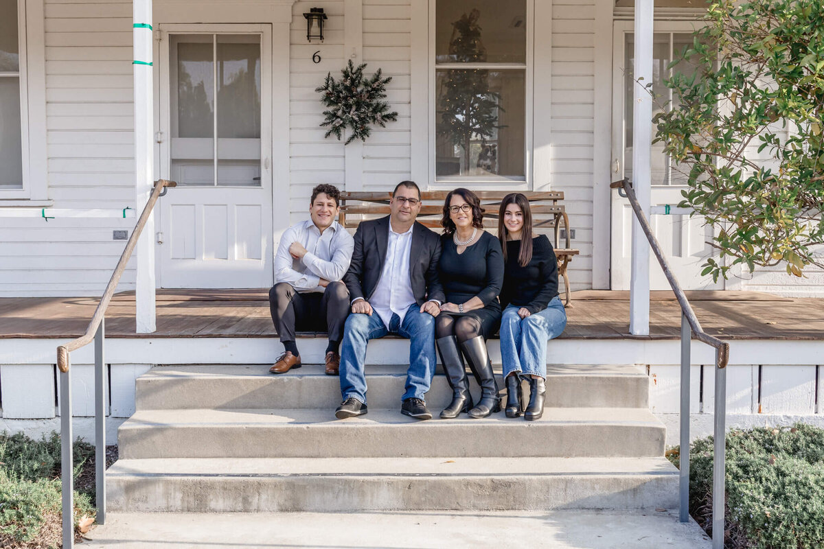 A family of four with dark hair wearing shades of blue and black looking at the camera from afar, sitting on the porch steps of an old, white and brown home at Katie Wheeler Ranch in Irvine.