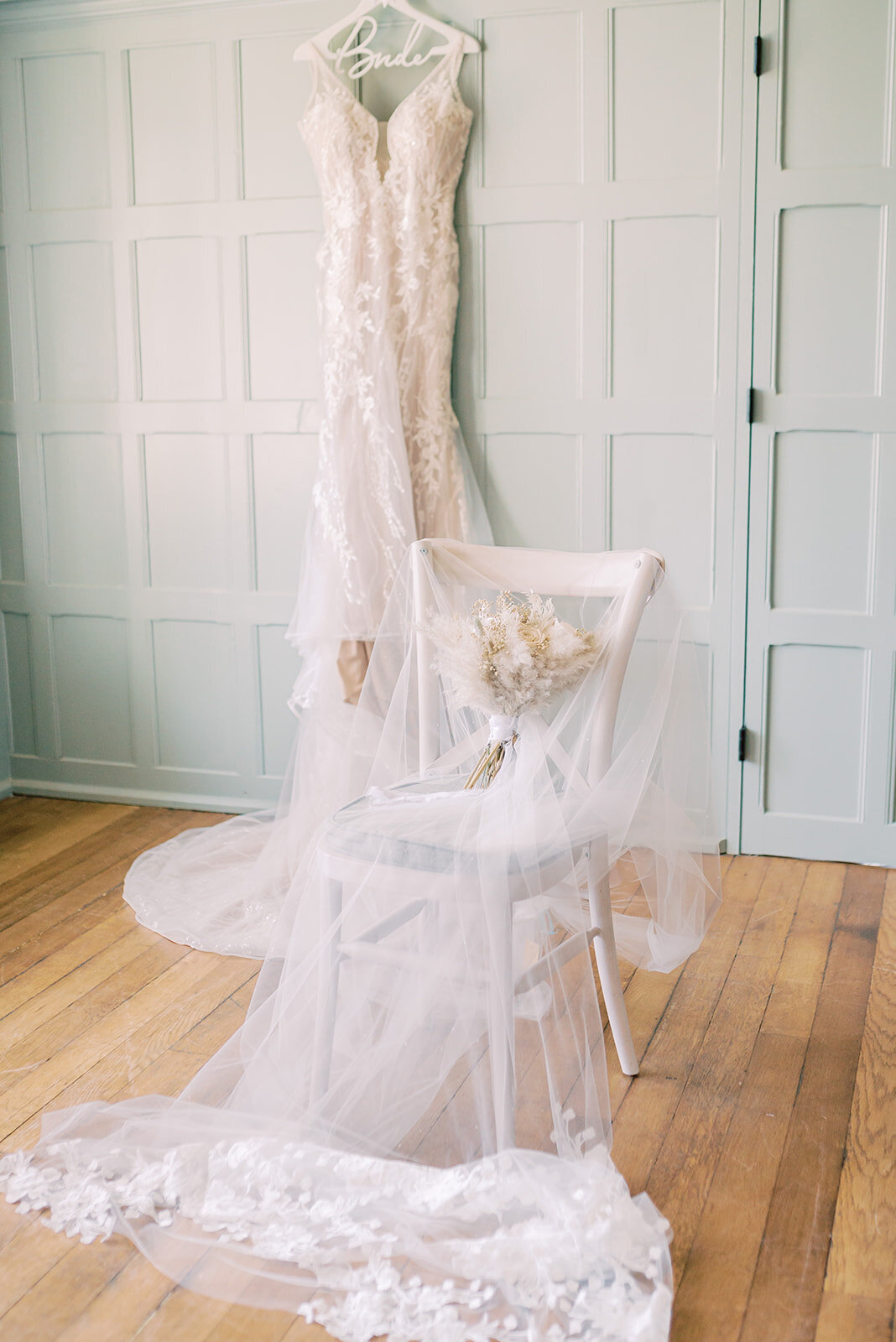 Light and airy image of the brides dress haging in the background and the veil and bouquet on a chair.