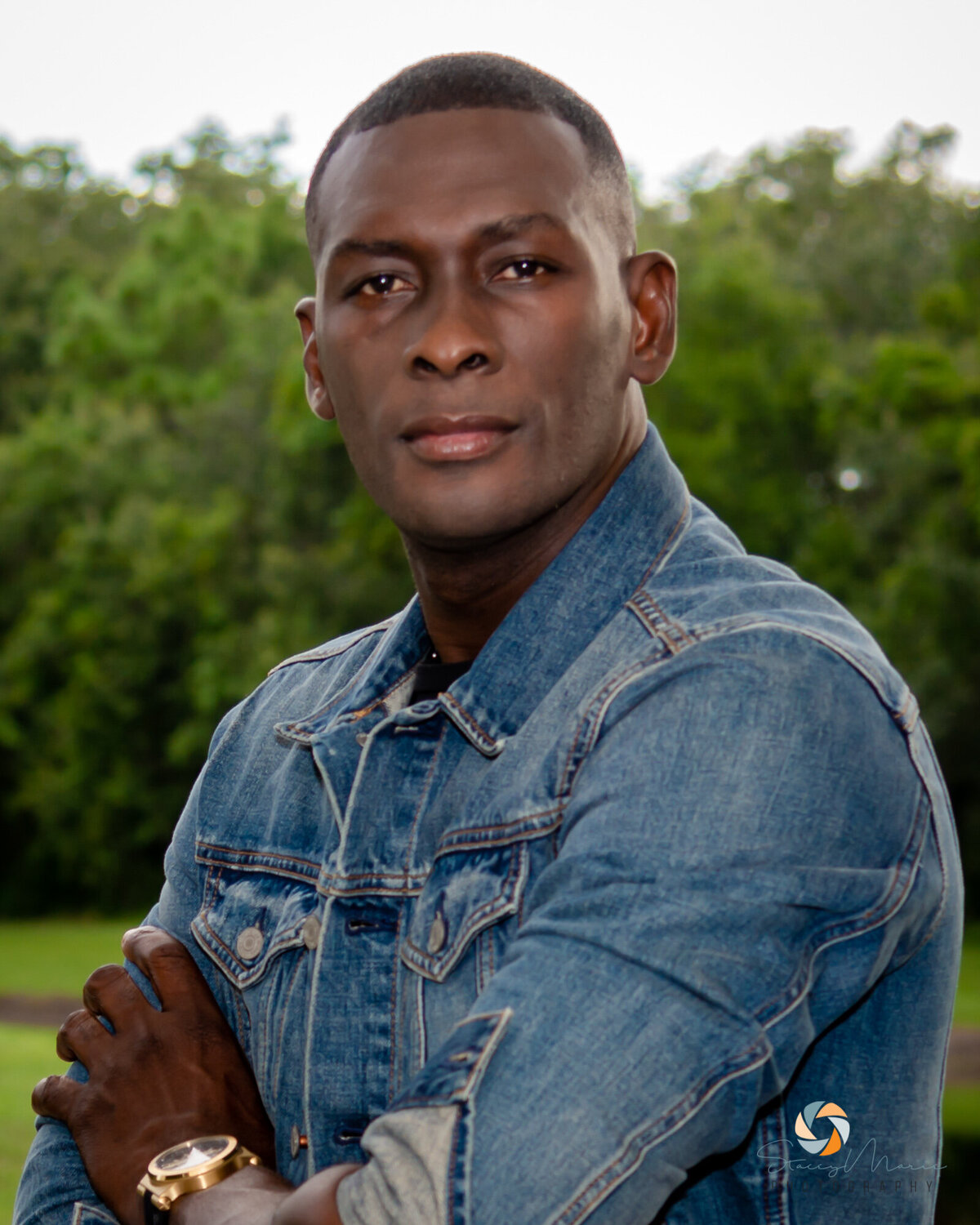 A man poses in a jean jacket in front of greenery.