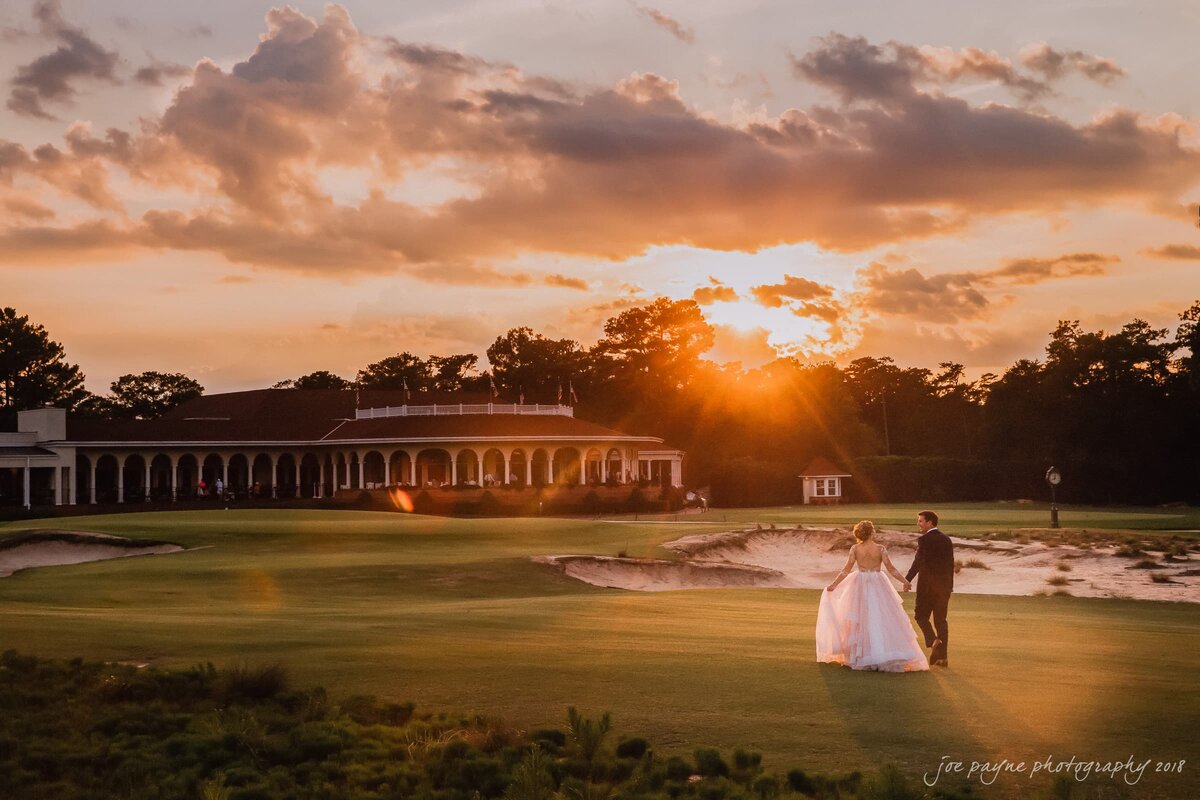 A wedding couple holding hands and walking on a golf course at sunset.