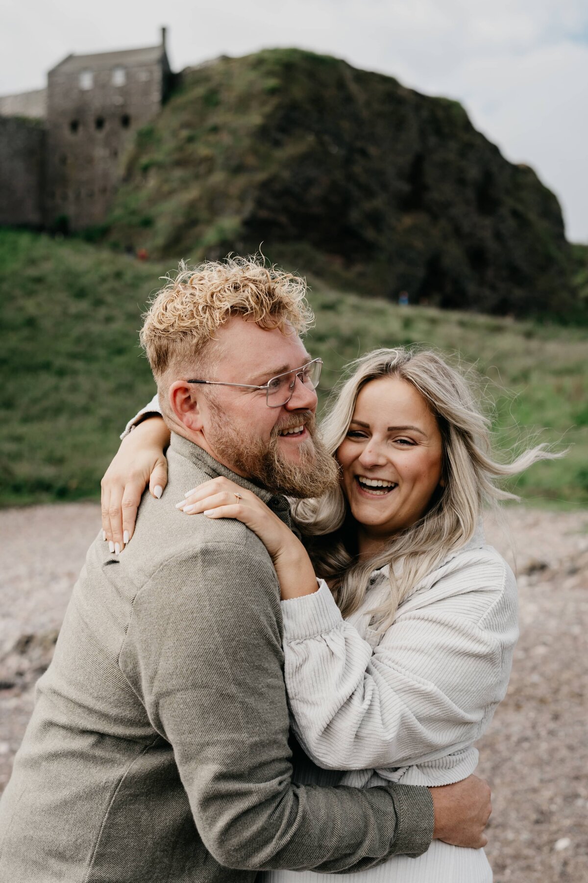 A couple laugh candidly with each other as they hug on the beach near Dunnottar Castle during their couple photoshoot.