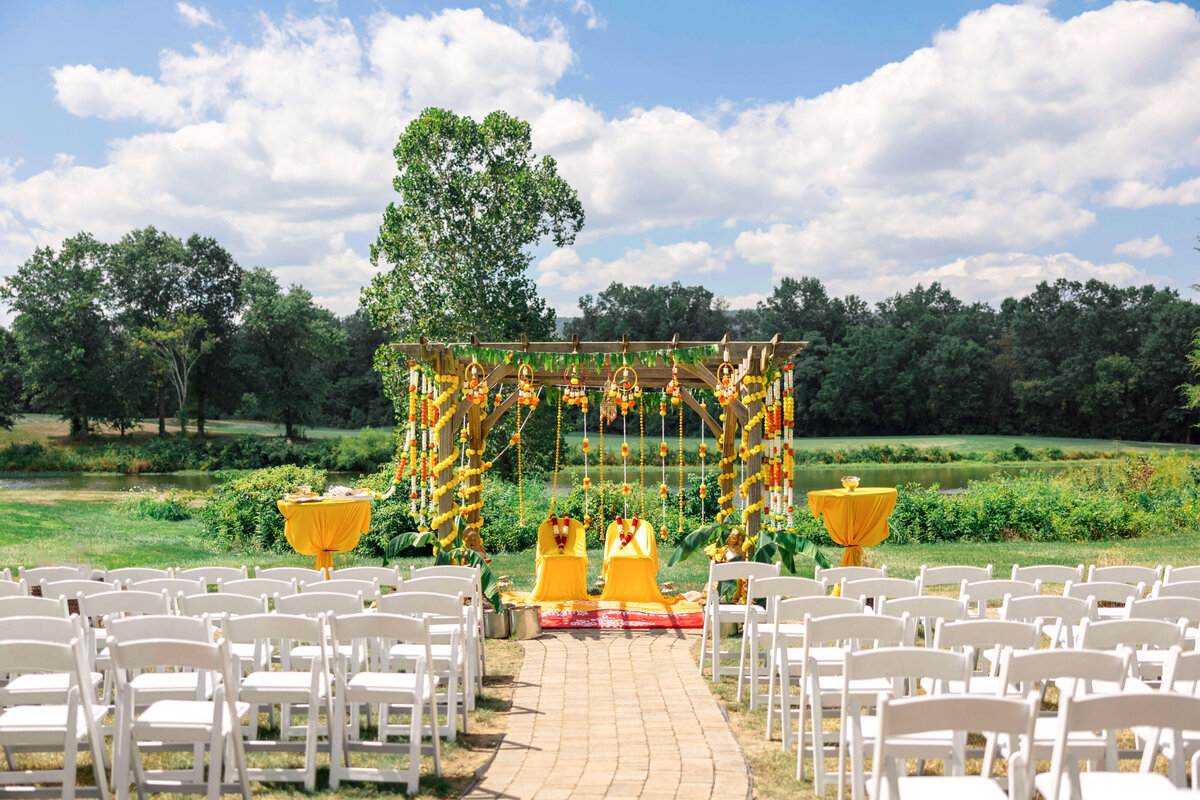 Outdoor wedding setup with a wooden mandap decorated with yellow and orange flowers. Two chairs are under the structure. Rows of white chairs are arranged on both sides of a path leading to the mandap, set against a lush green and tree-lined backdrop.