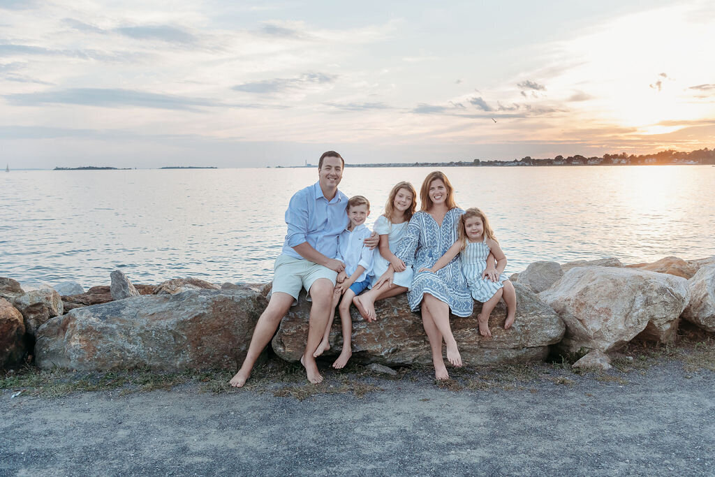 Connecticut family photographer captures family of five sitting on rocks during golden hour waterfront portrait session