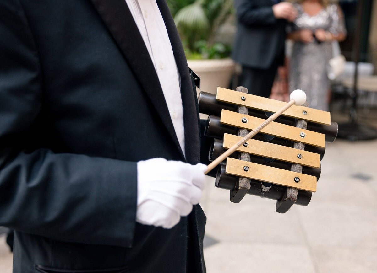 A person in a formal suit and white gloves holds a set of wooden tone blocks and a mallet. Blurred figures in the background suggest an elegant setting or event.