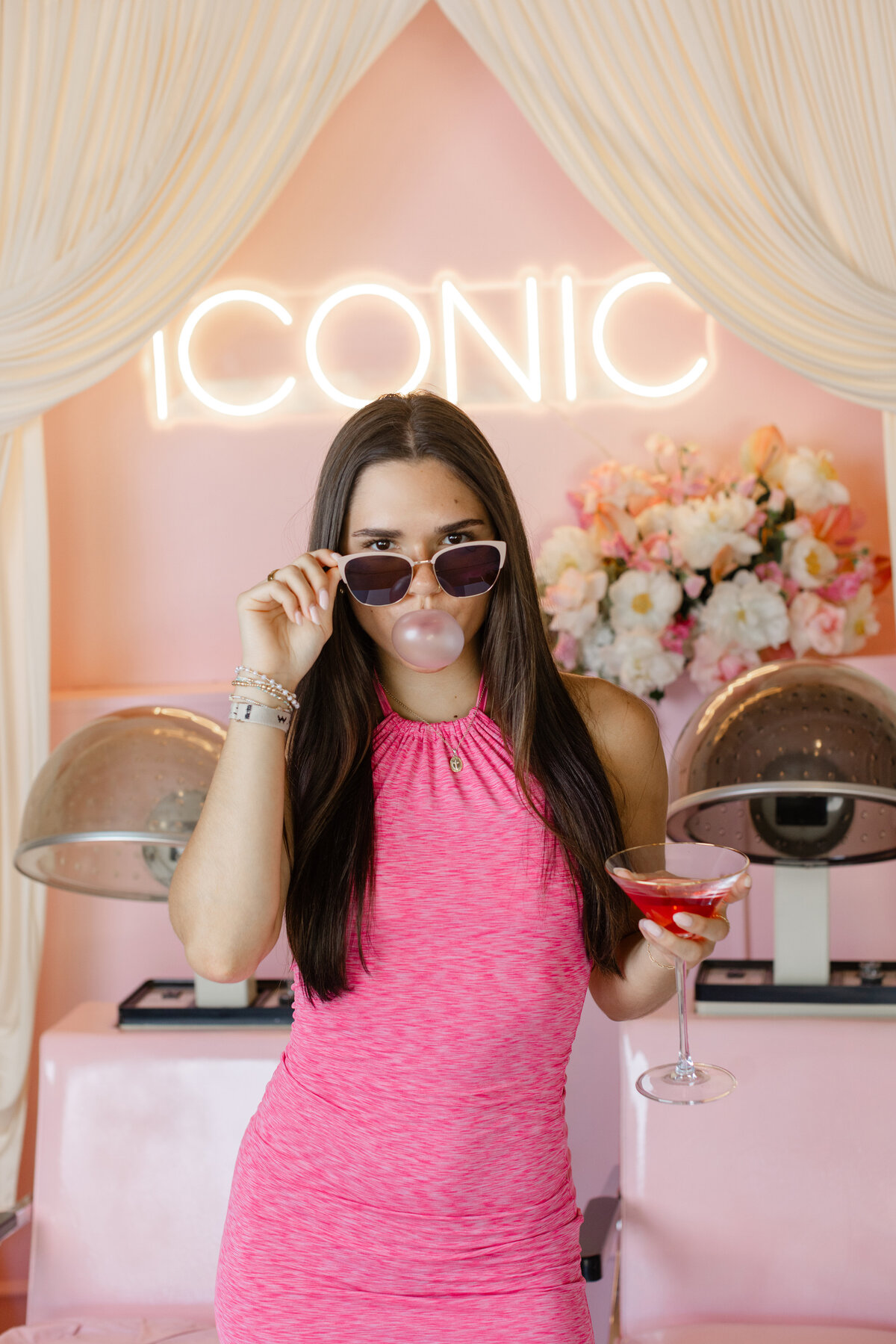 Greenville south Carolina branding photography shot of a woman with bubble gum and sunglasses with martini in a retro pink salon setup with disco balls and an "ICONIC" neon sign. Styled branding image by Dani Lacey.