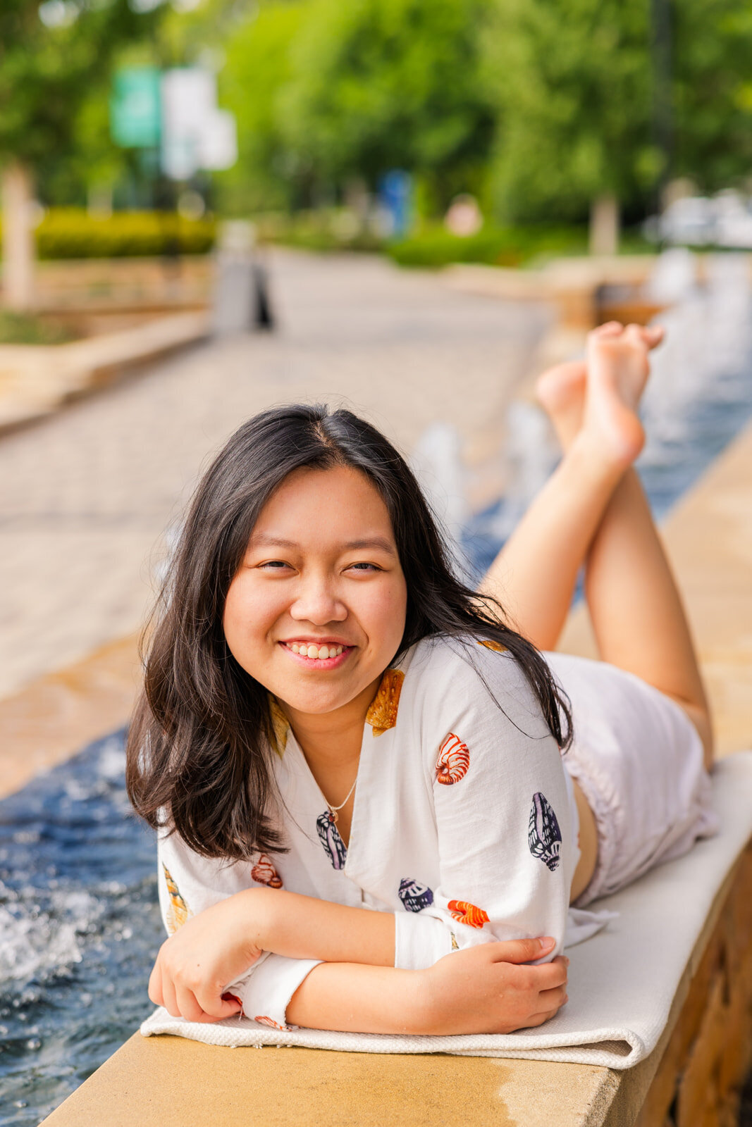 senior gilr laying down on her tummy arms crossed and feet up smiling at the camera in front of a fountain during photoshoot
