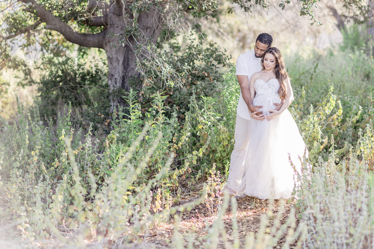 A pregnant woman and her husband in a green field cradling her belly with their heads together at Northwest Open Space