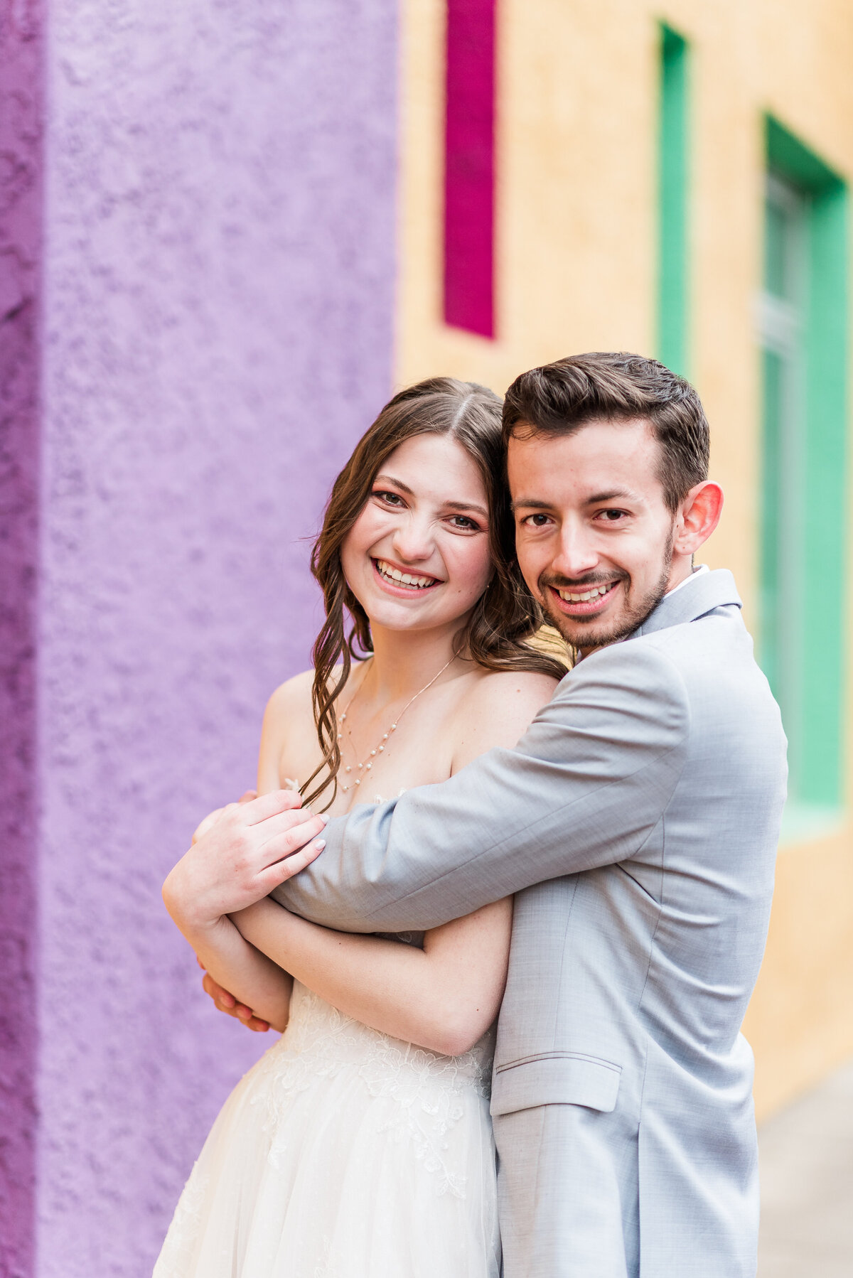 Groom in a light gray suit hugs his bride in her wedding dress from behind as they both smile toward the camera at Denver wedding photographer while standing in front of a purple, green, and yellow wall in downtown denver