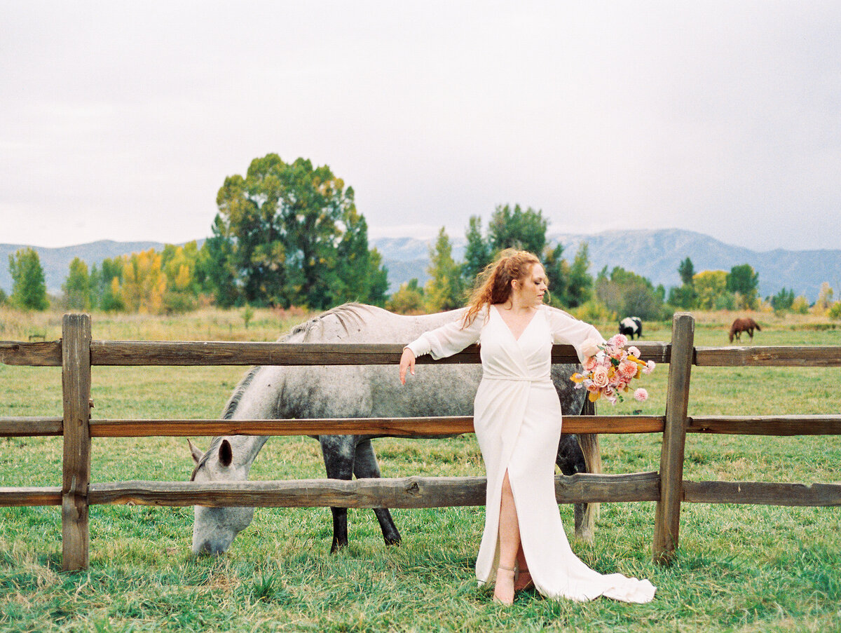Bride leans back on fence as the horse grazes on the background