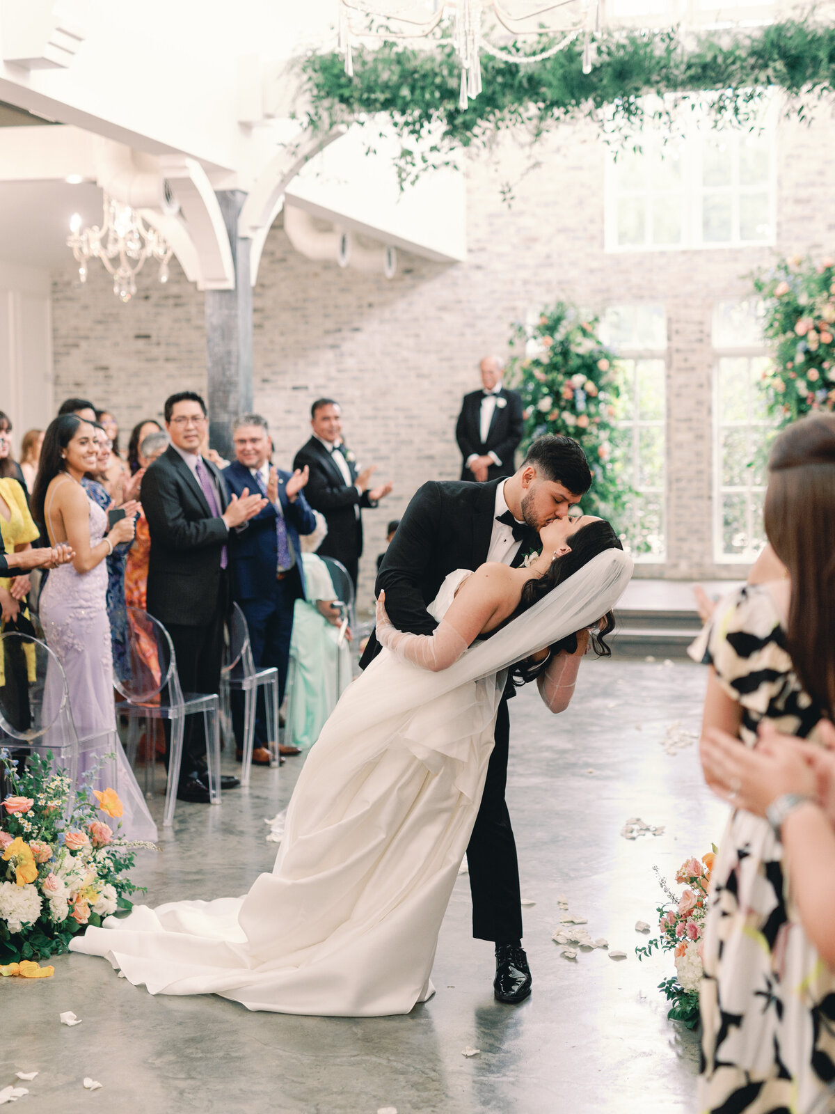 elegant bride with gloves and modern dress with bright and colorful garden party florals and groom in black suit walking down aisle during ceremony