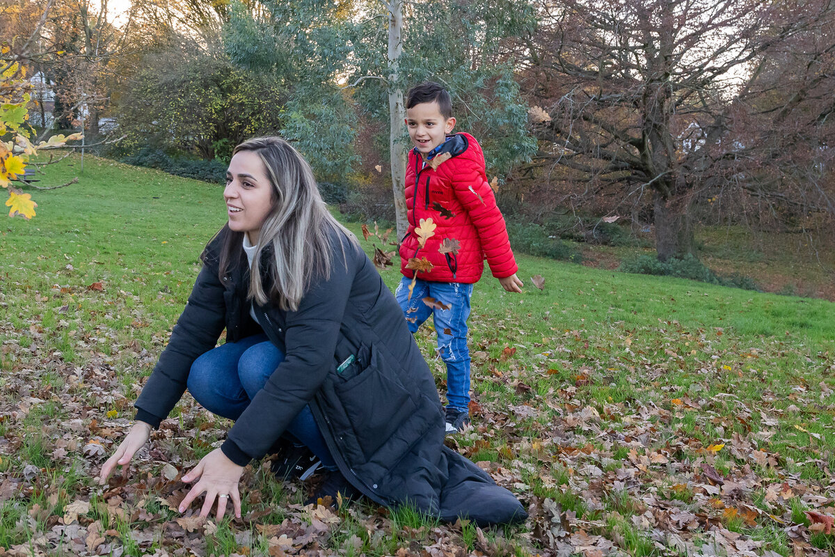 A woman and a young boy with a red jacket play with autumn leaves in a park, showing joyful expressions.