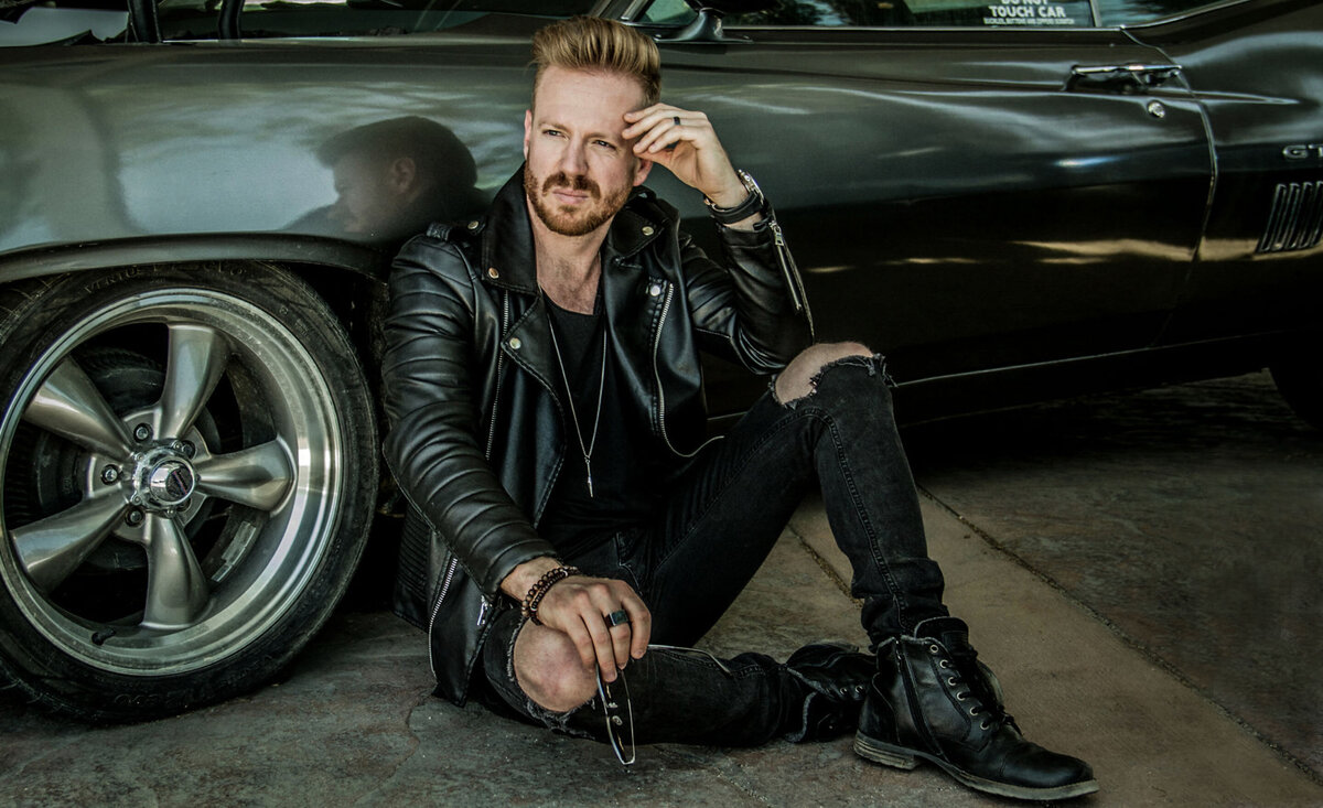 Male musician portrait Brad Saunders wearing black leather jacket black jeans sitting against old muscle car wheel