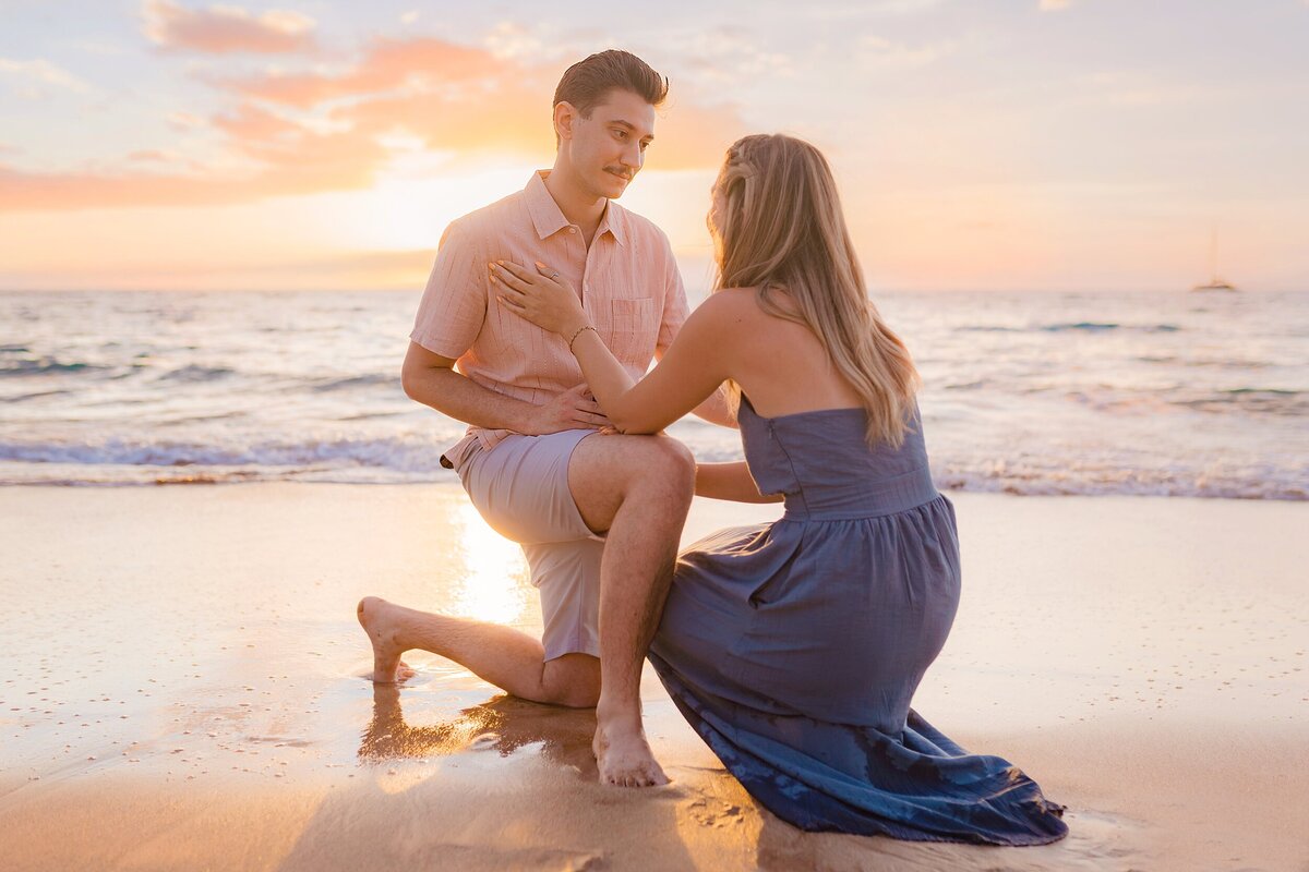 Blonde woman in a navy blue maxi dress accepts her partner's beach proposal in Maui at sunset