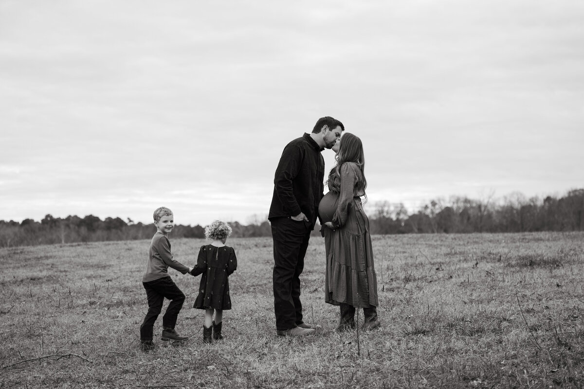 Black and white maternity photo of a family walking hand-in-hand through a field—capturing connection, anticipation, and timeless joy.