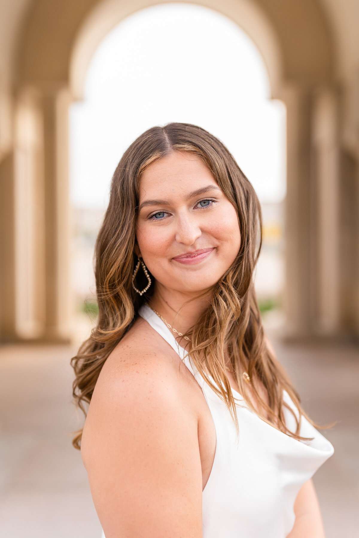 Texas A&M senior girl smiling over shoulder while wearing white dress in the archways of the Bell Tower