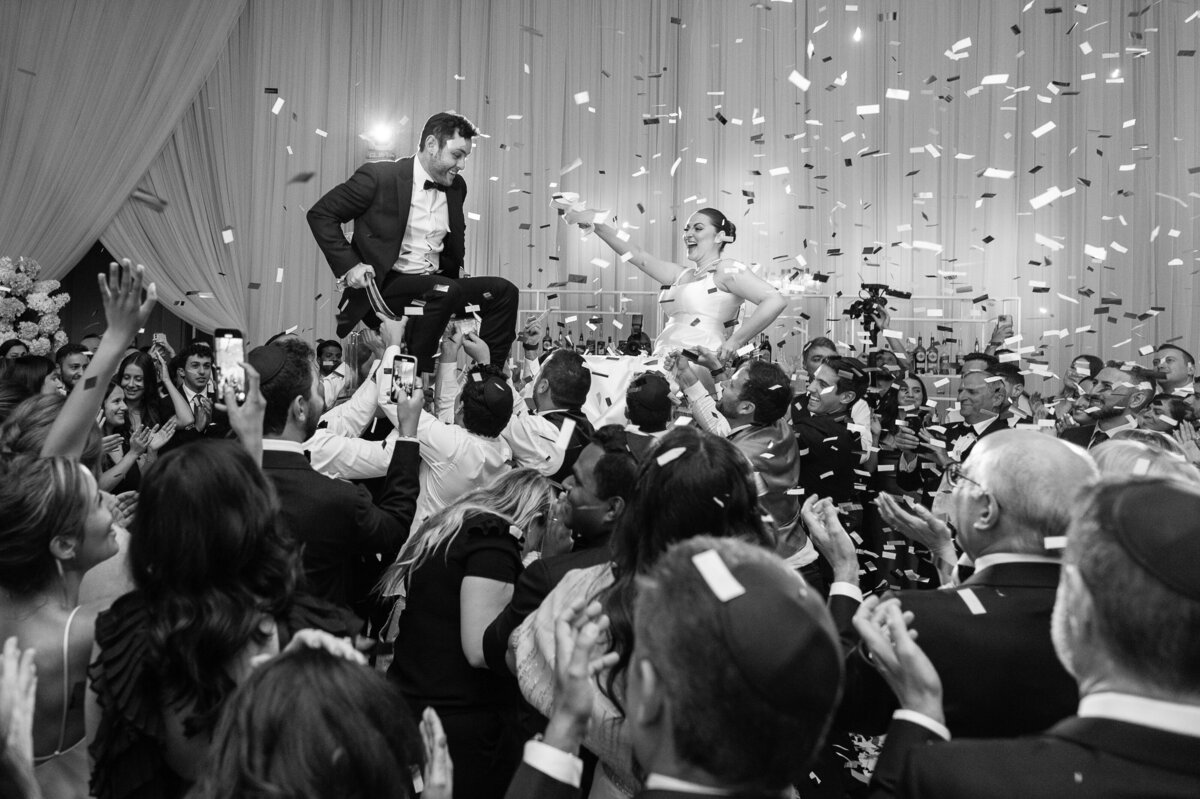 Black and white image of a couple on their wedding day being lifted on chairs during the hora. They are being showered with confetti.