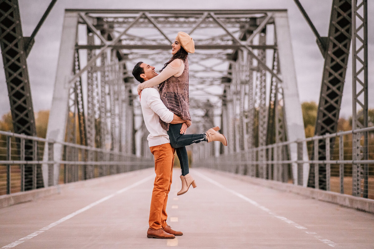 A man lifting a woman joyfully on a steel bridge, both smiling and gazing at each other lovingly.