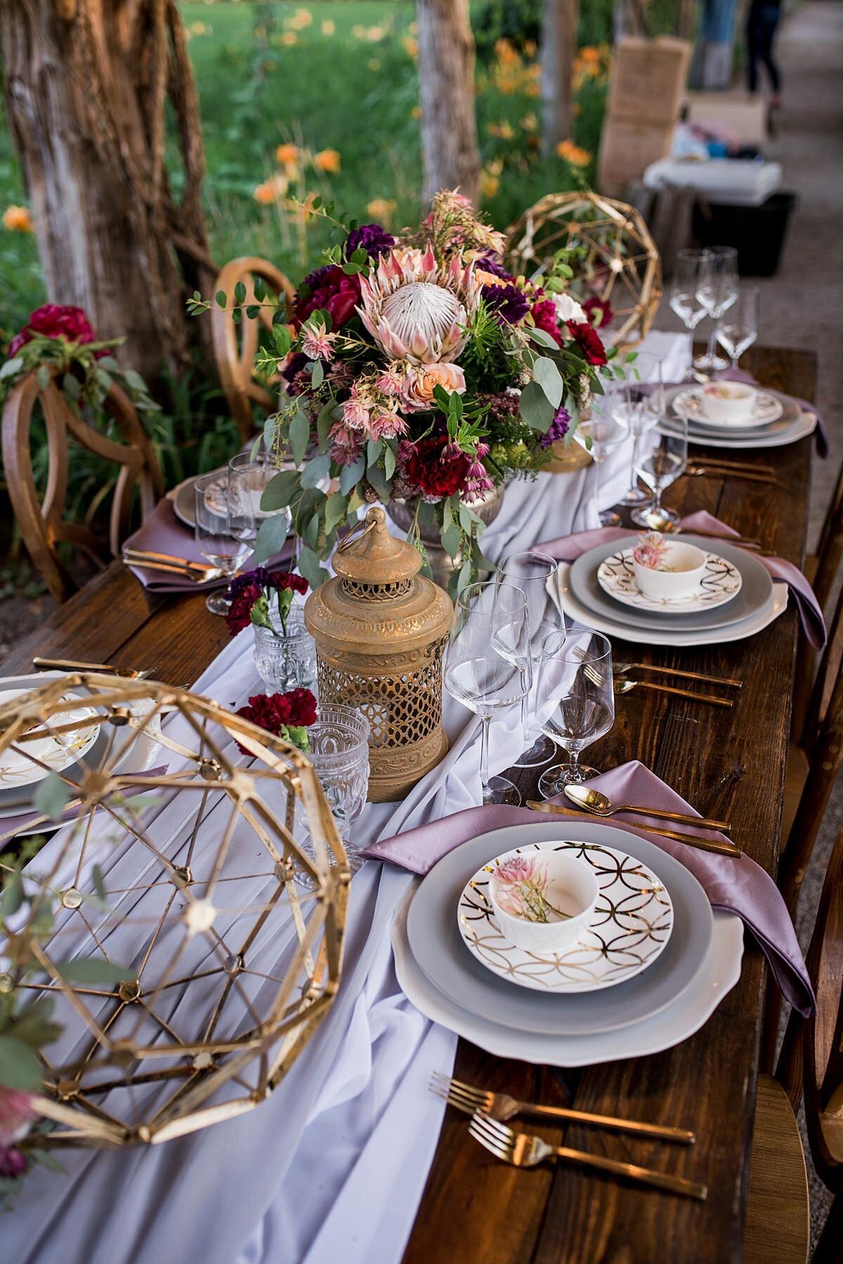 Dark wood farm table with soft gray organza table runner, gold moroccan lanterns and large gold geometric spheres with a large floral centerpiece. The centerpiece is in a gold footed bowl with king protea, burgundy dahlia, peach tea roses,  silver dollar eucalyptus and greenery. The table is set with gold flatware, a white charger with a scalloped edge, a gray dinner plate and a white salad plate with gold geometric detail. The plates are topped with a white bowl holding a prince protea with a purple napkin. The table is set in a lush garden under an arbor with wisteria.