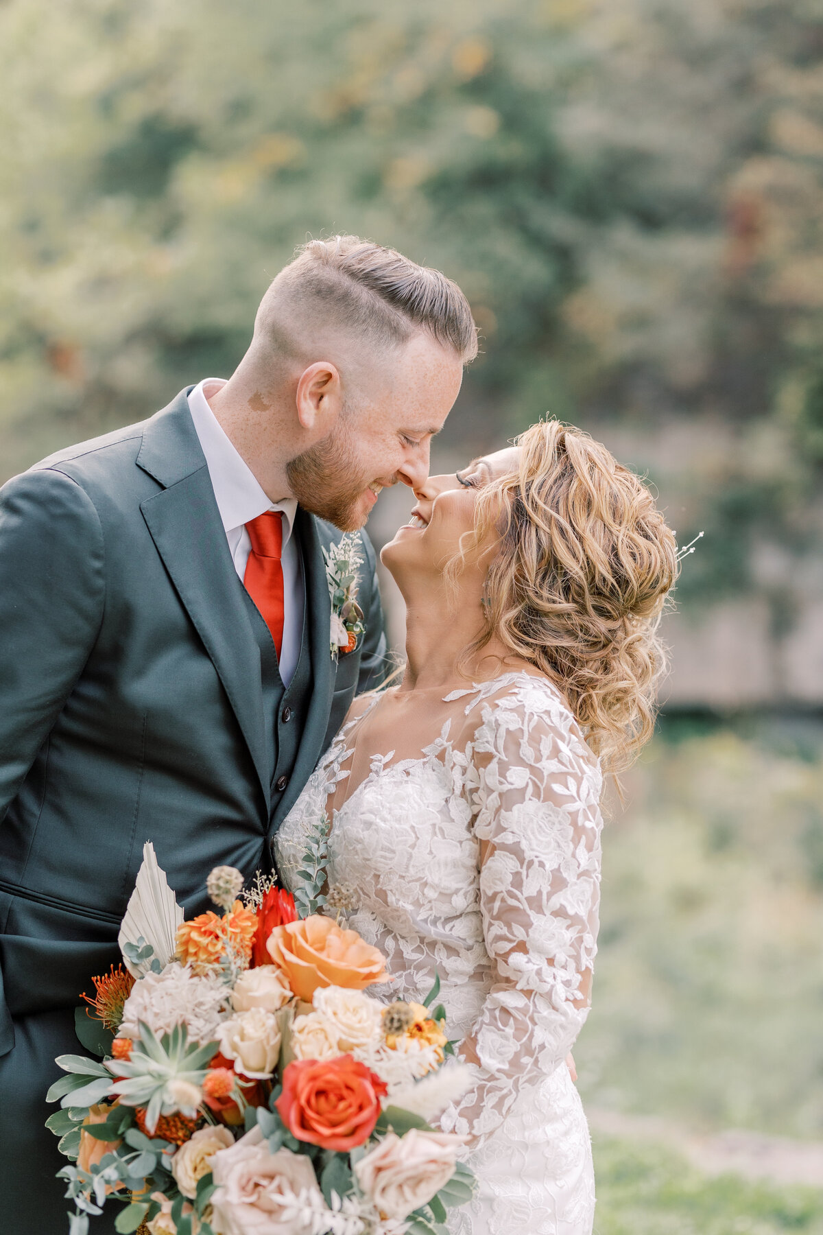A man in a green suit and a bride in a long sleeve lace gown press noses together while holding an orange, white, and cream bouquet with succulents on their wedding day.